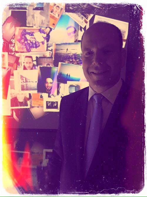 L'image présente un homme en costume avec une cravate, qui se tient devant un mur rempli de photographies. Les photos semblent évoquer divers souvenirs, probablement des moments importants et des lieux variés. L'éclairage donne une atmosphère chaleureuse et nostalgique à la scène, avec des teintes colorées qui ajoutent une dimension artistique. L'homme sourit, ce qui suggère un moment de convivialité ou de réflexion sur ces souvenirs représentés.