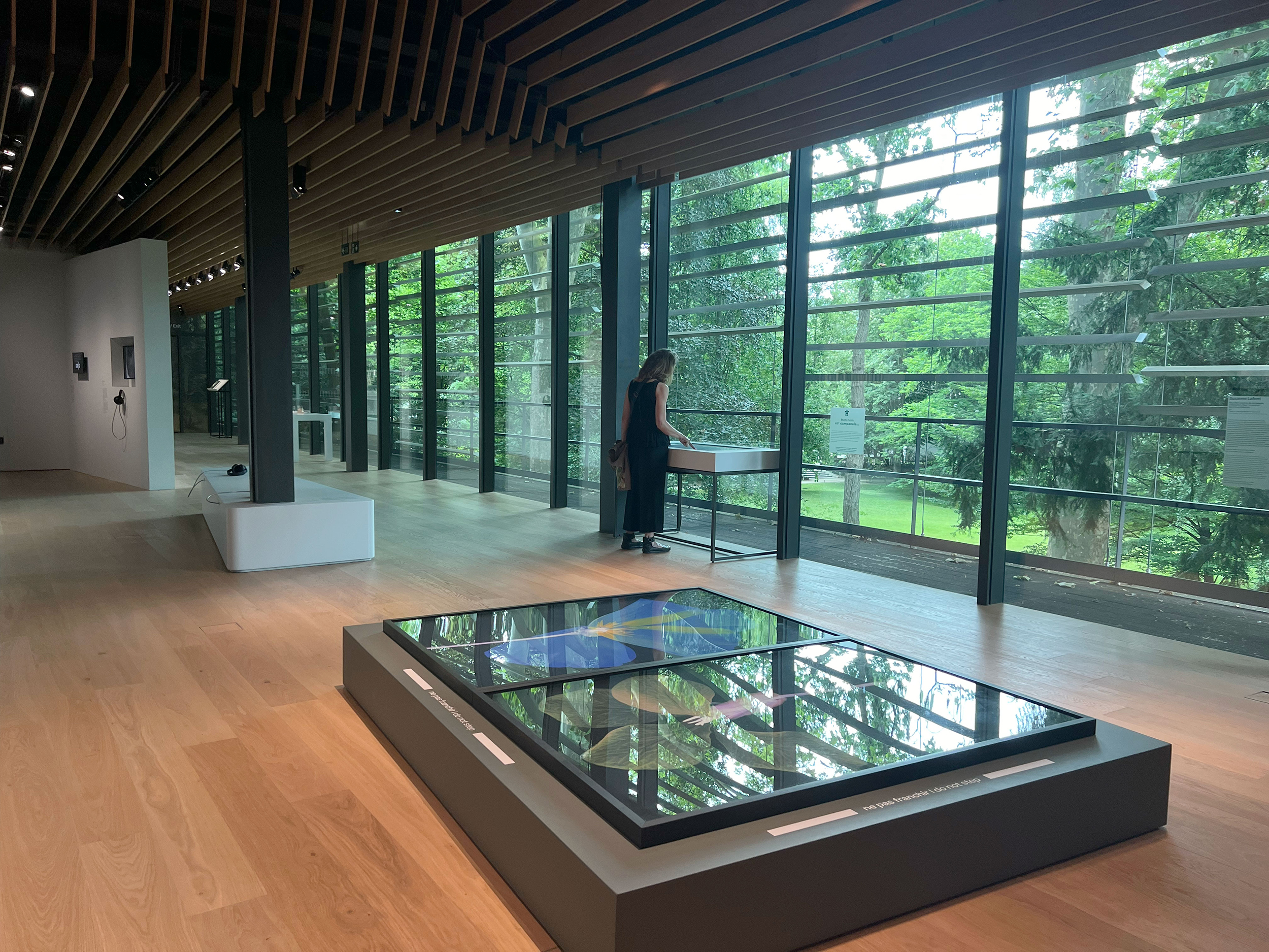 L'image montre l'intérieur d'un musée ou d'une galerie d'art, avec un sol en bois clair et de grandes fenêtres qui laissent entrer la lumière naturelle. À gauche, on aperçoit une personne debout près d'une table, semblant observer une œuvre d'art ou une exposition. Au centre, il y a une grande installation ou un tableau en verre qui présente des couleurs vives. L'environnement est verdoyant, offrant une vue sur l'extérieur avec des arbres et de la végétation. L'ensemble donne une impression de modernité et d'harmonie entre l'art et la nature.