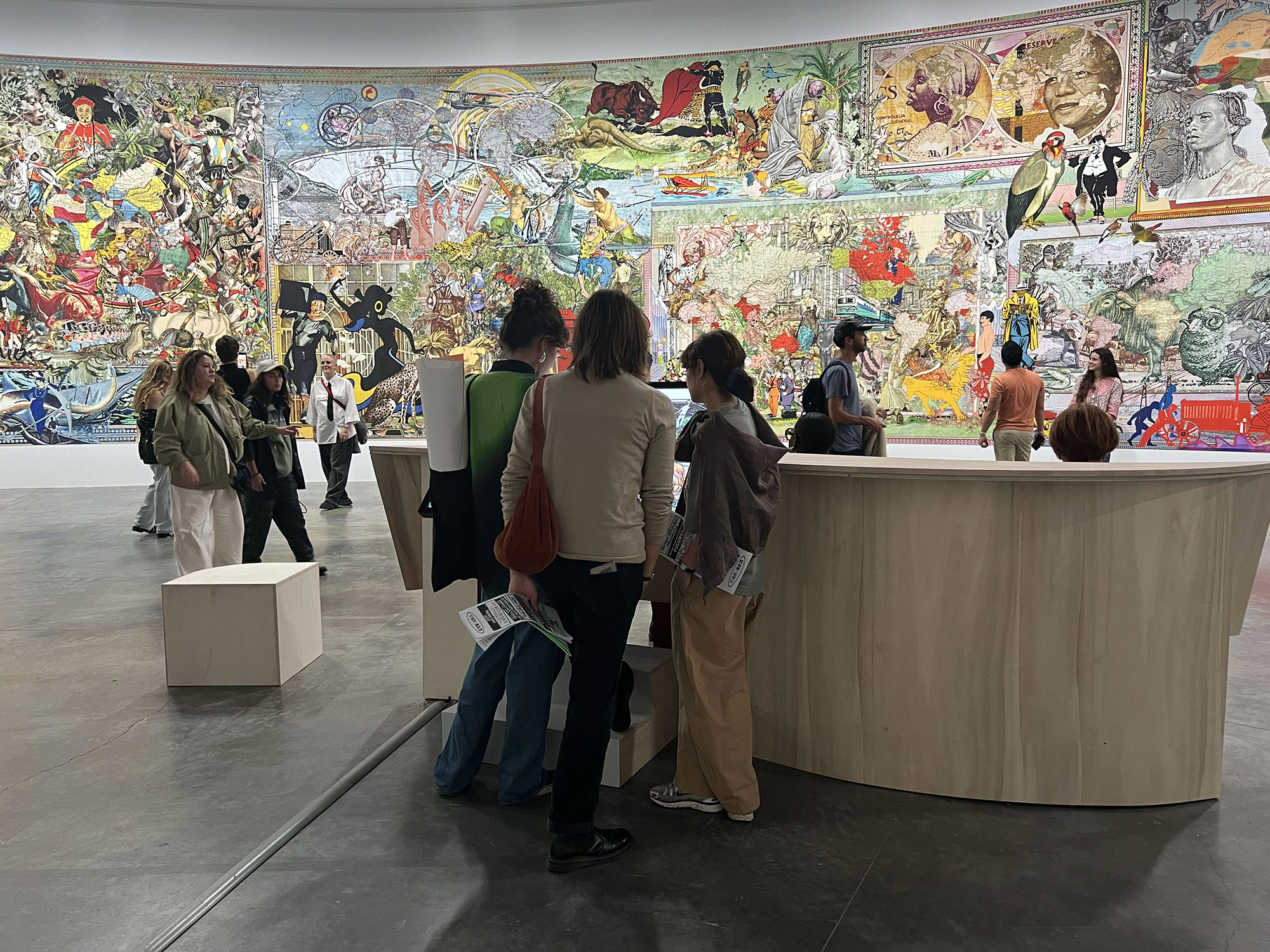 L'image montre une galerie d'art où plusieurs personnes explorent une œuvre murale colorée qui occupe toute la surface d'un mur. La scène est animée, avec des visiteurs discutant et observant l'art. Les personnages sur le mur semblent provenir de diverses influences artistiques et culturelles, créant une atmosphère dynamique et engageante. Des structures en bois, probablement des présentoirs ou des zones de repos, sont visibles au premier plan.