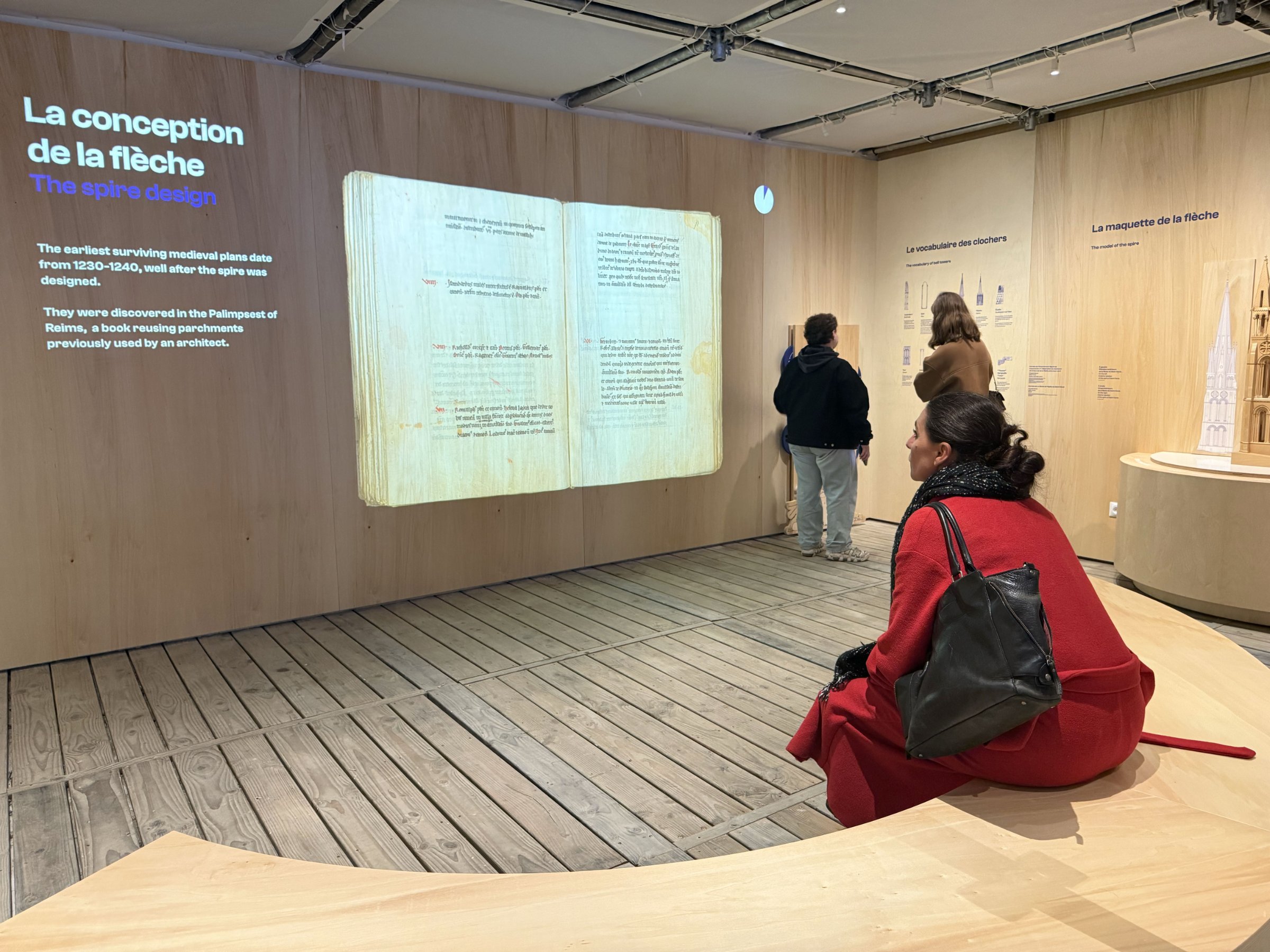 L'image montre une exposition dans un musée, où des visiteurs explorent des panneaux d'information. Au centre, on voit une femme assise sur une structure en bois, vêtue d'un manteau rouge et d'un sac à main noir. Les panneaux sur le mur incluent des informations sur le design des flèches architecturales, avec des illustrations et du texte en français. L'ambiance est moderne et épurée, avec des murs en bois et un sol en bois.