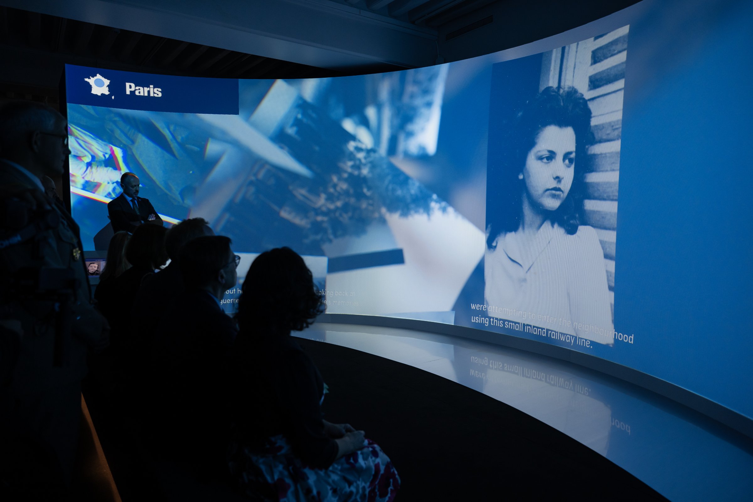 L'image montre une installation artistique ou un événement avec un écran incurvé affichant des contenus visuels. À gauche, une personne semble présenter ou parler, tandis qu'à droite, on voit une photo en noir et blanc d'une femme. Le fond est illuminé et les spectateurs, assis en face, regardent attentivement. Le mot « Paris » est affiché en haut. L'atmosphère semble être celle d'une exposition ou d'une présentation multimédia.