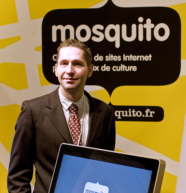 L'image montre un homme en costume, debout devant un fond jaune avec le mot "mosquito" en lettres blanches. Il semble souriant et confiant, et il y a un ordinateur portable ou un écran devant lui qui affiche également le logo ou le nom "mosquito". L'ensemble donne une impression professionnelle, probablement en lien avec une présentation ou un événement.