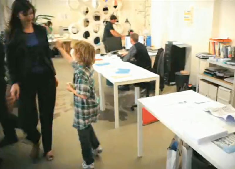 L'image montre un bureau avec plusieurs personnes. À l'avant, une femme en tenue professionnelle se tient près d'un enfant, qui semble lui faire un geste. L'enfant porte une chemise à carreaux. À l'arrière-plan, on peut voir d'autres personnes en train de travailler, ainsi que des meubles de bureau tels que des bureaux blancs et des étagères. L'environnement semble moderne et créatif, avec des éléments de décoration murale.