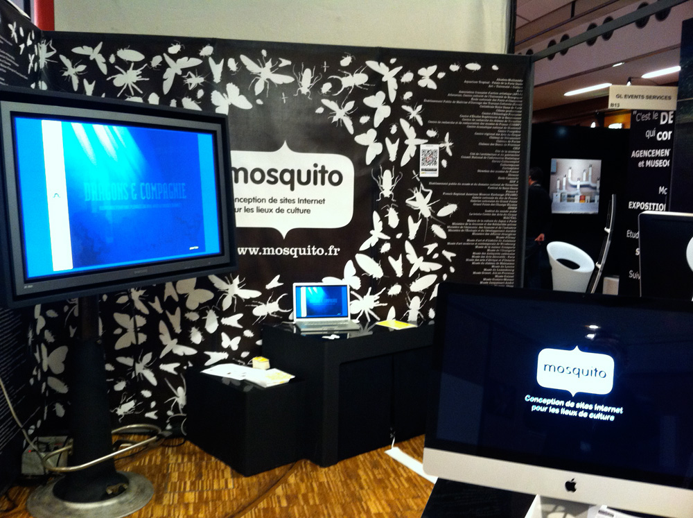 L'image montre un stand d'exposition avec un fond décoré de motifs de papillons. On y trouve un écran plat monté sur un support, affichant une présentation, ainsi qu'un ordinateur portable et un autre écran compact sur une table noire. Le logo de "mosquito" est visible sur l'un des écrans. Le stand semble être dédié à la conception de sites Internet pour des projets culturels.