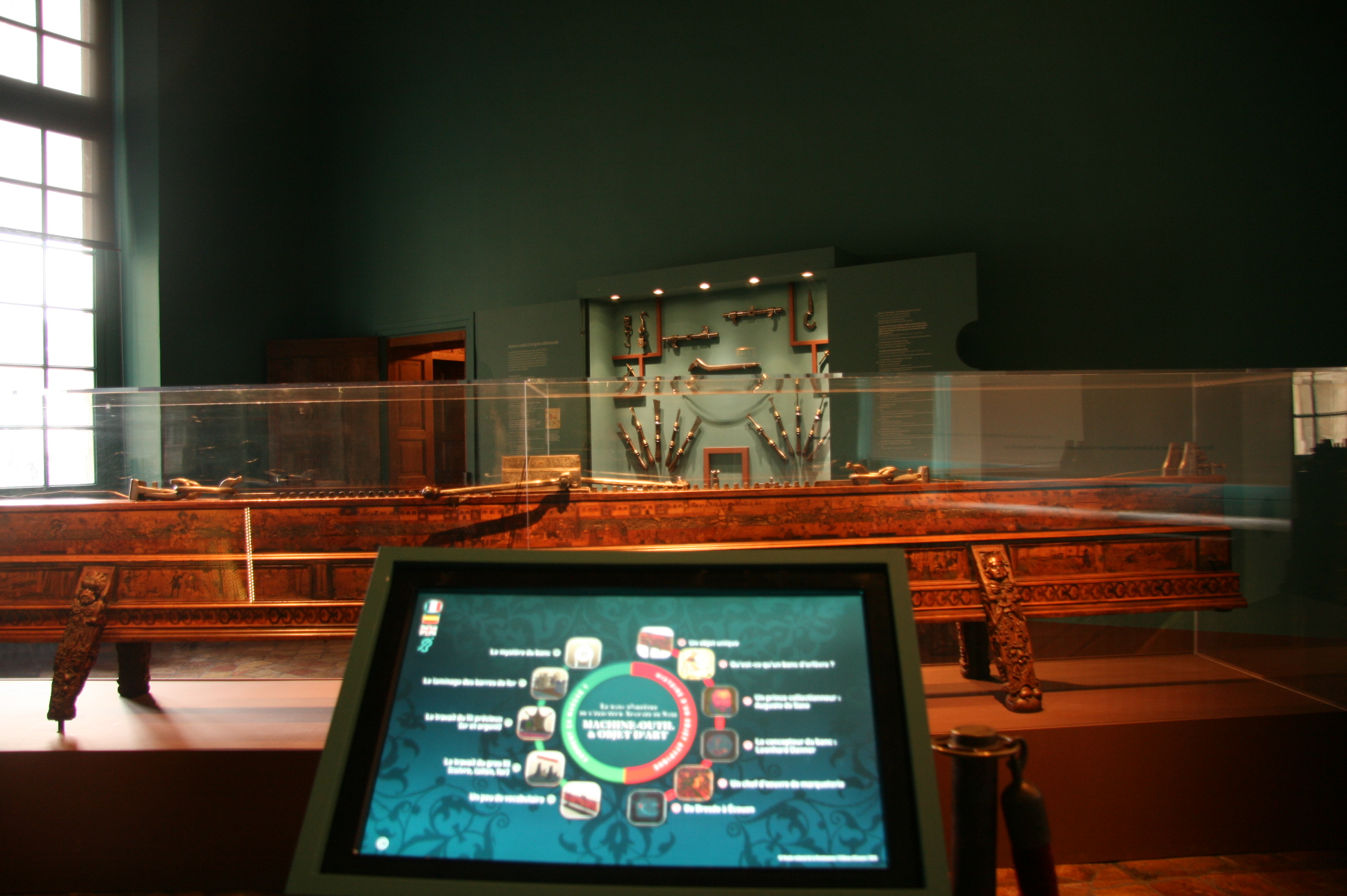 L'image montre un intérieur de musée où l'on peut voir un grand meuble en bois décoré, probablement une pièce d'artisanat ou un objet historique exposé dans une vitrine transparente. À l'arrière-plan, un mur est décoré avec plusieurs objets accrochés, qui pourraient être des armes ou des outils anciens. Au premier plan, il y a un écran interactif qui semble fournir des informations sur l'exposition. L'ambiance est calme, avec des murs peints dans une teinte sombre qui met en valeur les objets exposés.