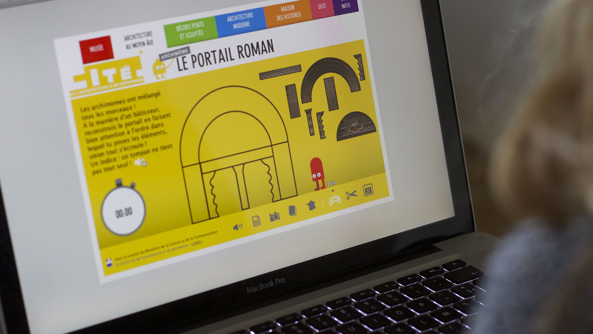 L'image montre une personne utilisant un ordinateur portable, probablement un MacBook. L'écran affiche une interface colorée, centrée sur un thème d'architecture romane. On peut y voir un grand arc, des motifs géométriques et des éléments graphiques sur fond jaune. Un chronomètre est visible, indiquant un compte à rebours de 00:00. Cela semble être une leçon ou une activité interactive sur les caractéristiques de l'architecture romane.