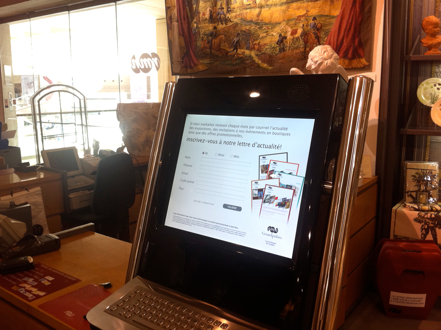 L'image montre un kiosque informatique moderne placé sur un comptoir. L'écran tactile est affiché, avec un formulaire pour s'inscrire à une lettre d'actualité, indiquant où entrer des informations. On aperçoit également des objets en arrière-plan, tels que des vitrines et une tapisserie. La conception du kiosque semble élégante et contemporaine.