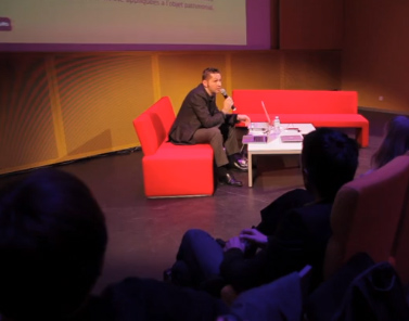 L'image montre un événement en intérieur où un intervenant est assis sur un canapé rouge, tenant un microphone et parlant à un public. Il semble concentré sur son discours, tandis que des membres du public, dont on voit les silhouettes, l'écoutent attentivement. L'ambiance est moderne et professionnelle, avec un éclairage doux.