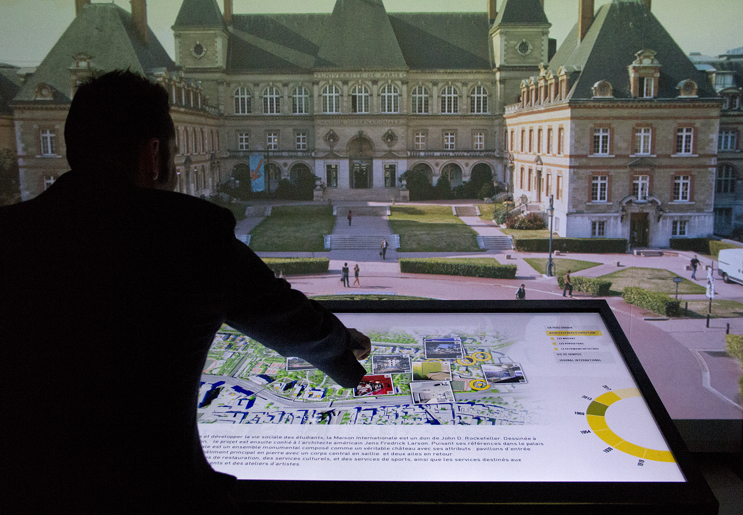 L'image montre une personne, probablement un homme, se tenant devant un grand écran qui affiche une vue de la place autour d'un bâtiment historique. Il interagit avec une carte affichée sur une surface tactile, pointant du doigt un endroit spécifique. Le fond révèle une architecture classique avec des détails élégants et une atmosphère de découverte urbaine. L'ensemble donne une impression de modernité alliée à l'histoire, comme une exposition ou un projet d'aménagement.
