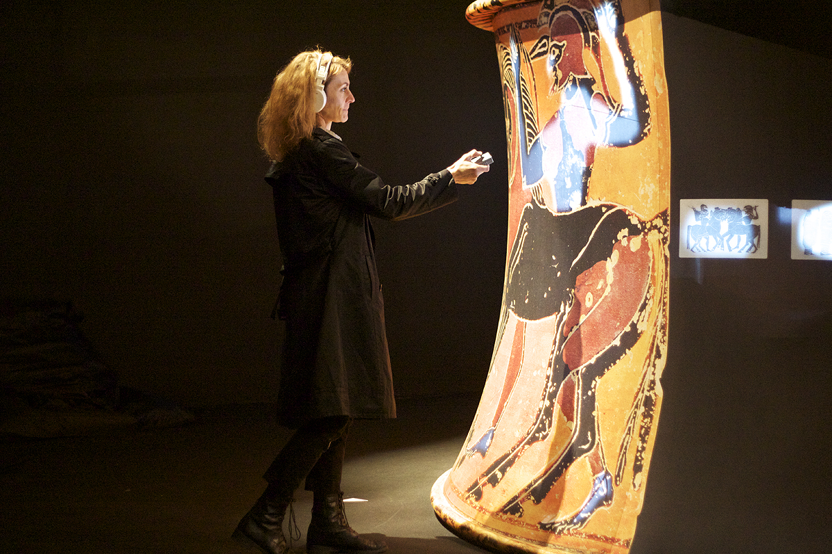 L'image montre une femme en train d'interagir avec un objet d'art, probablement une grande jarre ou un vase antique. Celui-ci est décoré de peintures représentant des figures humaines ou des scènes. La femme, vêtue d'un manteau noir, semble fasciné par la représentation sur le vase, ce qui crée une ambiance d'exploration et d'appréciation de l'art historique dans un cadre muséal ou d'exposition. L'éclairage met en valeur les détails des peintures, soulignant leur importance.