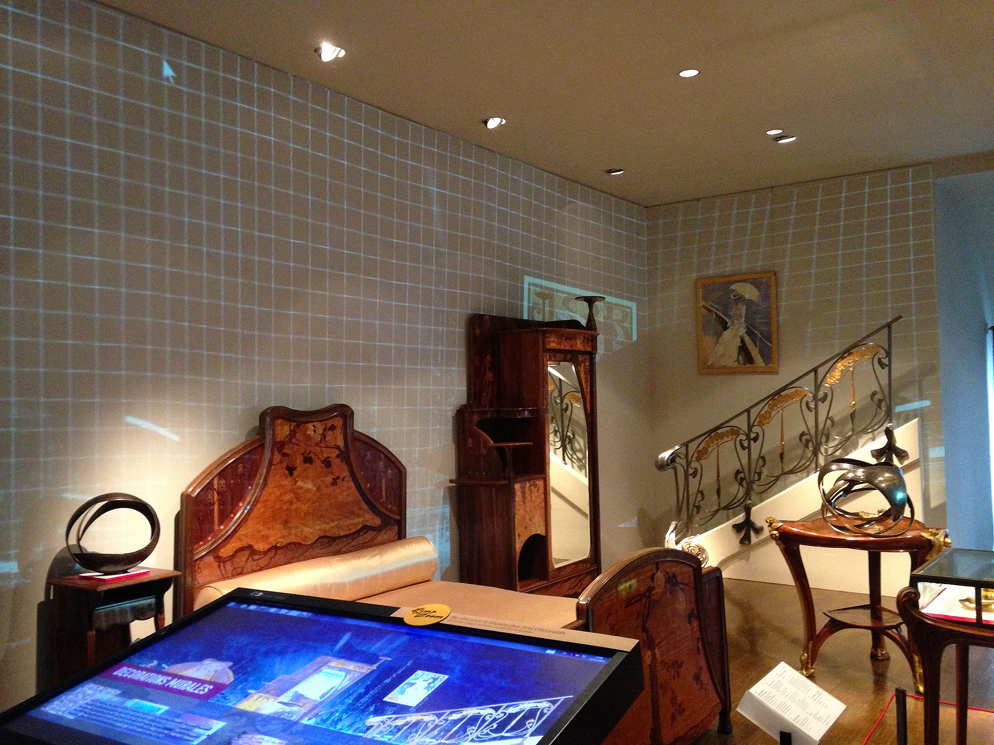 L'image montre une pièce aménagée de manière élégante, avec des meubles en bois sculpté. On peut voir un canapé et une chaise ornés, ainsi qu'un paravent. Une décoration murale projetée crée un effet de grille sur le mur, ajoutant une touche moderne à l'ensemble. Un escalier avec une rampe décorative se trouve à l'arrière-plan, tandis qu'un écran interactif est placé au premier plan, probablement utilisé pour fournir des informations sur les objets exposés. L'éclairage est doux et met en valeur le design des meubles.