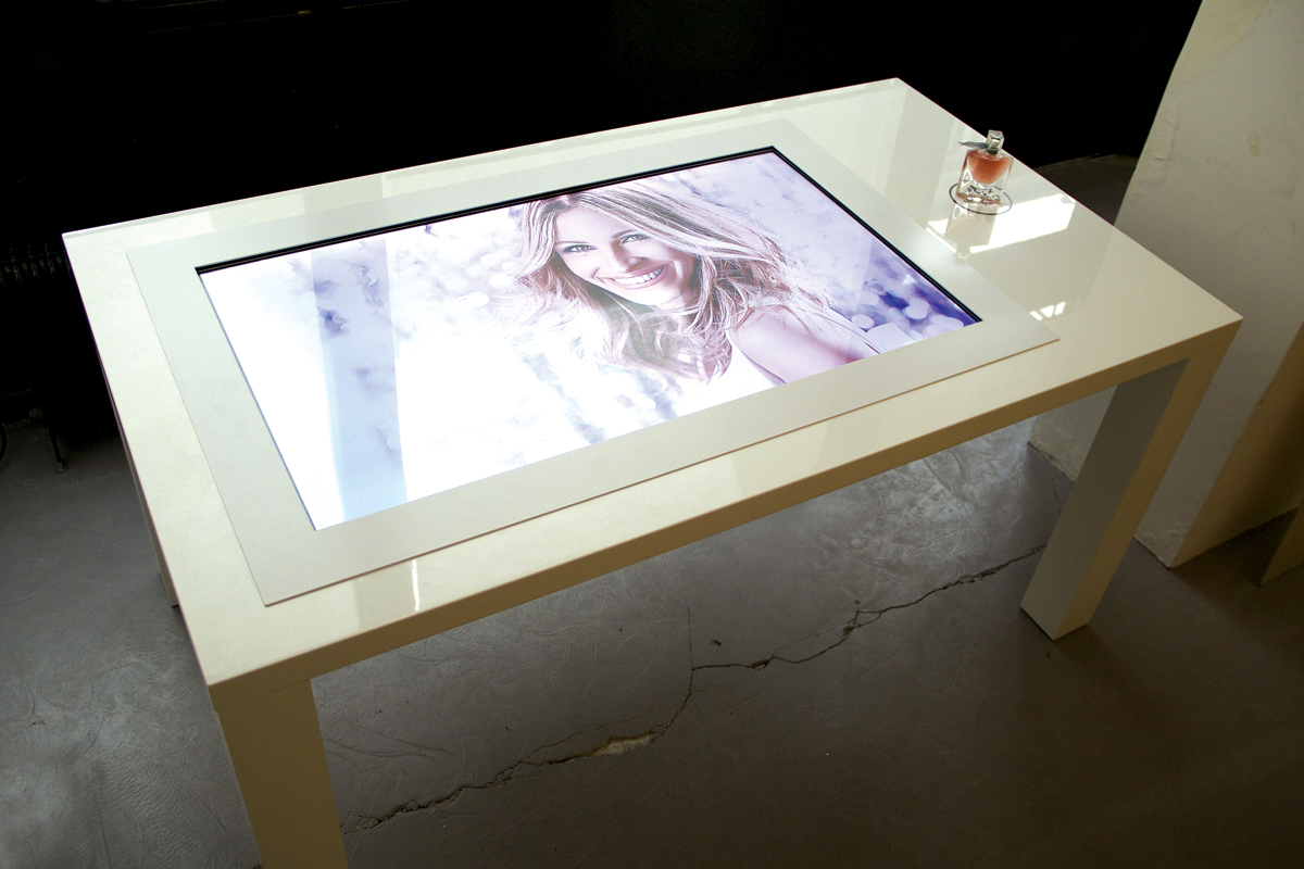 L'image montre une table moderne avec un plateau en verre. Sur ce plateau se trouve un écran tactile affichant une image, probablement d'une personne souriante. À côté de l'écran, il y a un flacon de parfum élégamment placé. La table a un design contemporain, avec des lignes épurées et une finition blanche, et elle est située dans un environnement intérieur minimaliste.