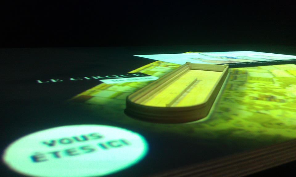 L'image montre une surface sur laquelle se trouve un livre ouvert, avec un accent sur le titre "Le Cirque". À côté, il y a un objet allongé, qui semble être un modèle ou une maquette, et une projection indique "VOUS ÊTES ICI !", suggérant une sorte d'interaction ou d'orientation dans un espace donné. L'ensemble est éclairé de manière à créer une atmosphère immersive et interactive.