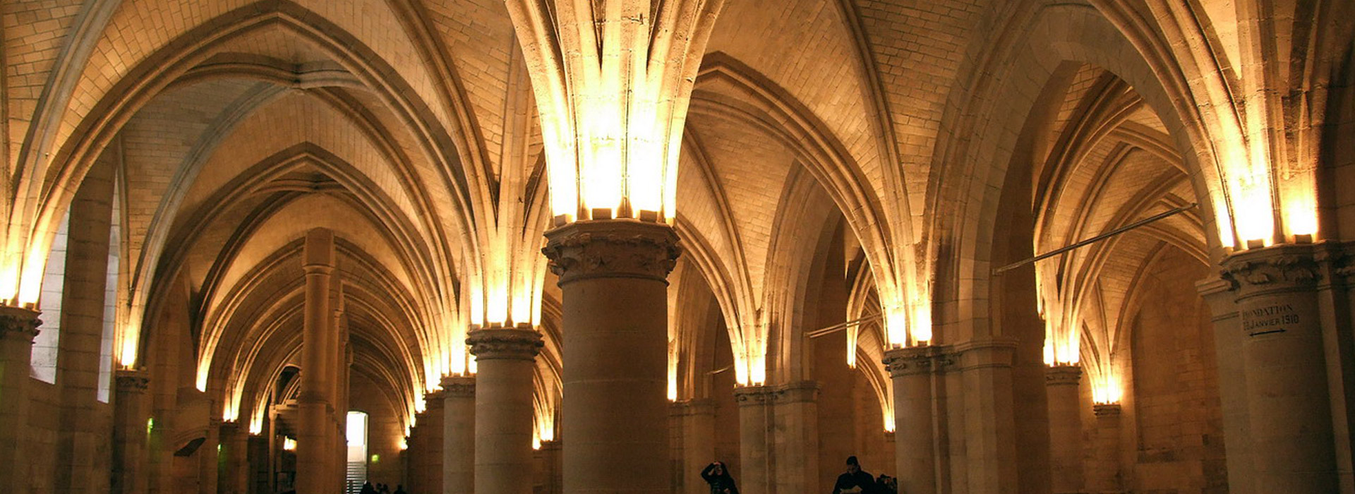 L'image montre un intérieur architectural majestueux, probablement une voûte d'un bâtiment historique. Les arches en pierre, avec des détails raffinés, s'élèvent vers le plafond, créant une atmosphère impressionnante. La lumière douce qui illumine les colonnes et les murs ajoute une ambiance chaleureuse et accueillante. On peut voir des silhouettes de personnes se déplaçant dans cet espace, soulignant la grandeur et la beauté de l'architecture.