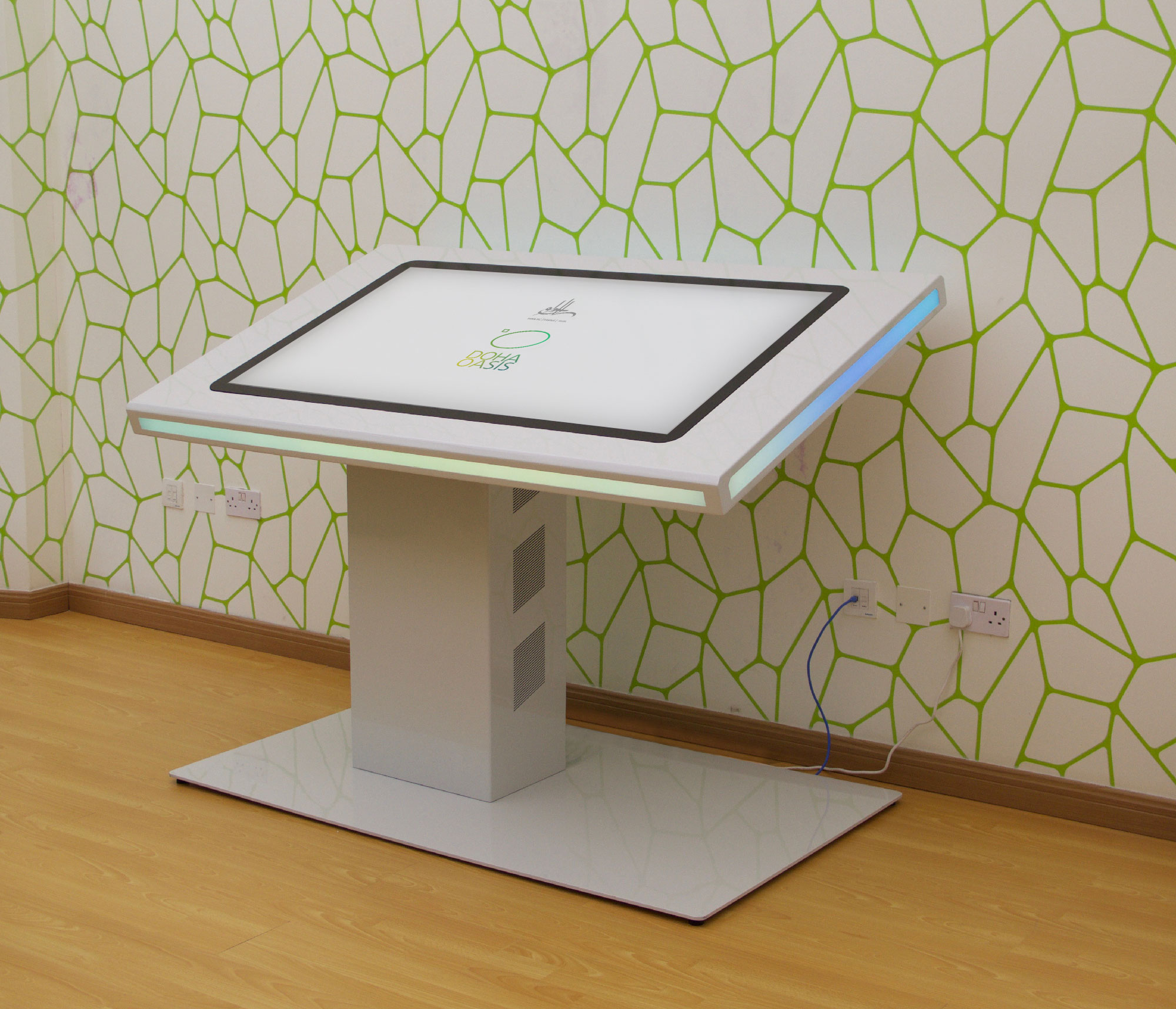 L'image montre une table interactive moderne qui semble conçue pour des utilisations technologiques ou éducatives. Elle est rectangulaire et inclinée, avec un écran tactile sur le dessus. La base de la table est en métal brillant, de couleur blanche, et elle repose sur un socle large. Le fond est orné d'un motif géométrique vert sur un mur clair, ajoutant une touche contemporaine à l'environnement. On peut également voir des prises électriques à proximité de la table.