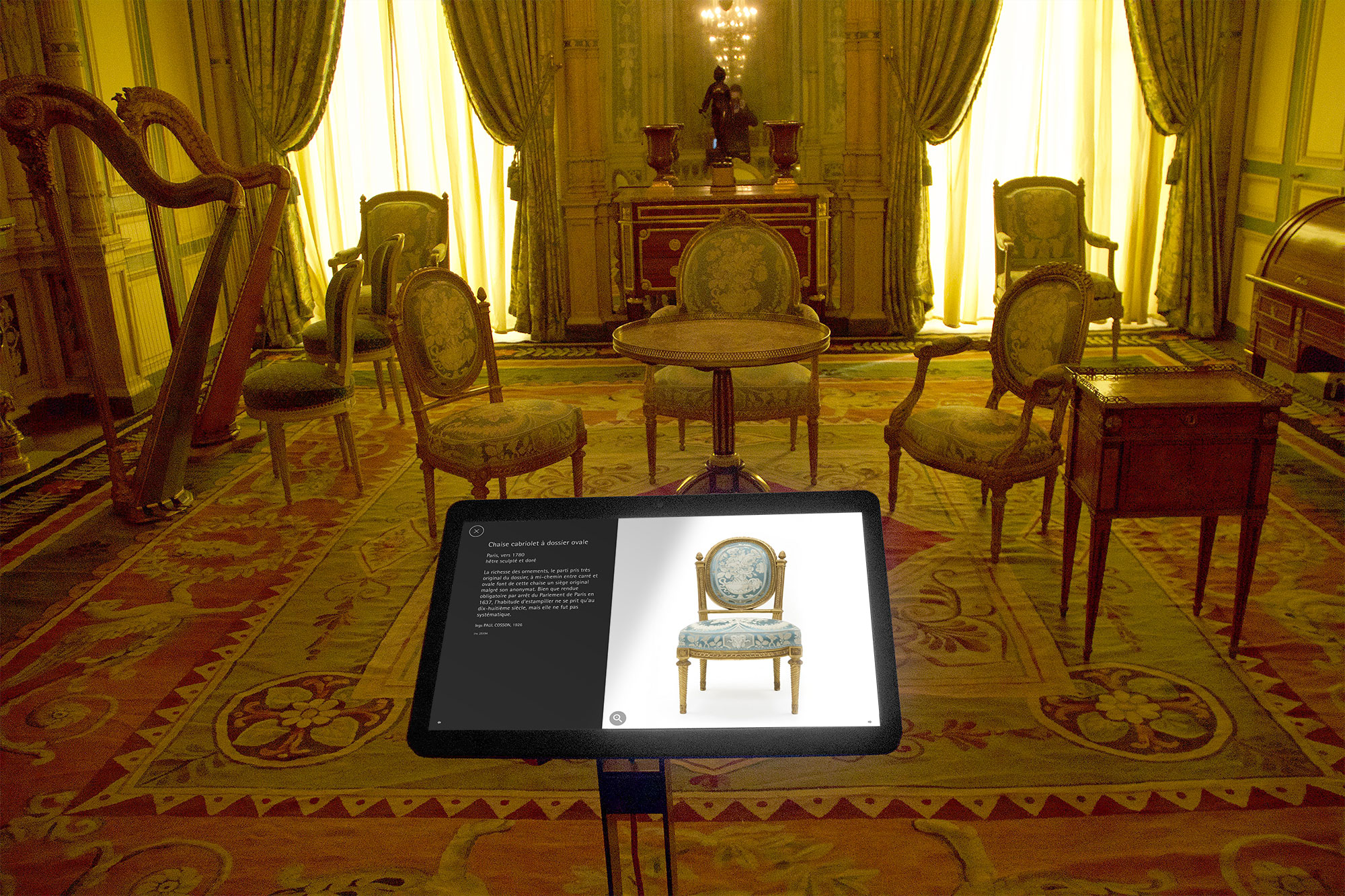 L'image montre une pièce élégante, apparemment un salon, avec plusieurs chaises ornées et une petite table en bois. Les rideaux sont tirés, laissant entrer une lumière douce qui met en valeur les détails de la décoration. Au centre, un écran interactif affiche des informations sur une chaise, ajoutant une touche moderne à cet espace traditionnel. Le sol est recouvert d'un tapis richement décoré, renforçant l'ambiance classique de la pièce.