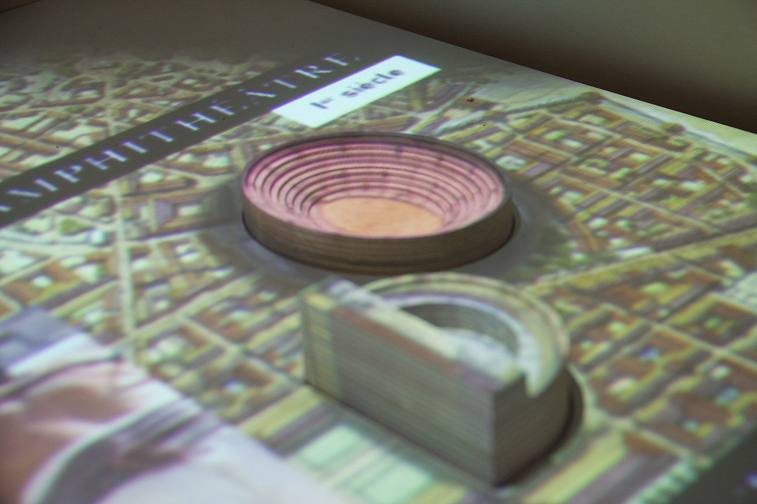 L'image montre une projection d'un amphithéâtre, probablement romain, avec des représentations en 3D. On peut voir des couches concentriques qui illustrent l'intérieur de l'amphithéâtre, ainsi qu'une vue d'ensemble de la zone environnante. La projection semble être sur une surface plane, donnant une idée claire de la structure et de son emplacement dans le contexte urbain.