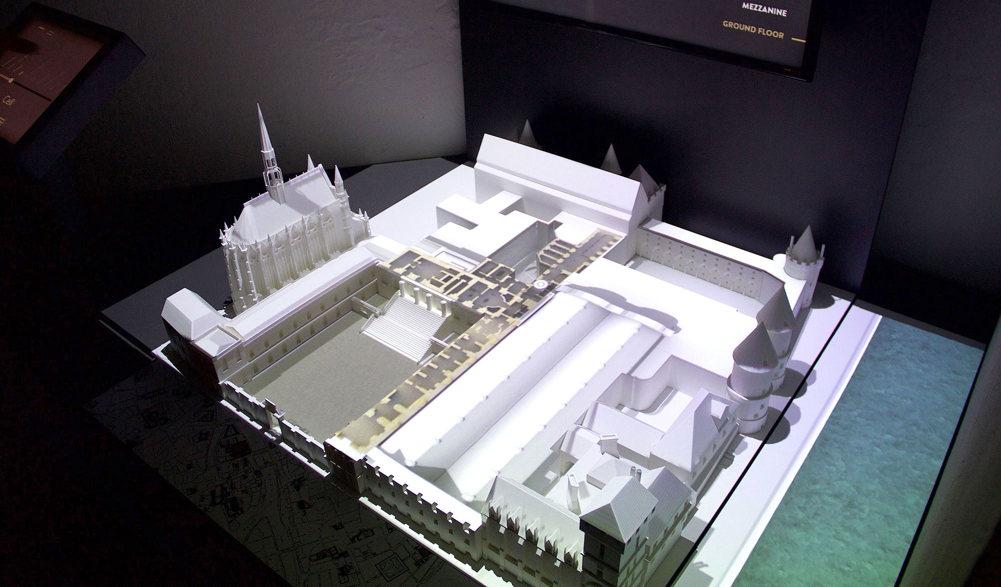 L'image montre une maquette architecturale en trois dimensions, probablement d'un bâtiment ou d'un site historique. La maquette est éclairée, mettant en évidence divers détails, comme les formes des bâtiments, les toits et les espaces environnants. On peut distinguer des structures avec des éléments architecturaux variés, et il semble qu'il y ait une partie en hauteur, probablement une tour. L'illustration permet de visualiser l'agencement des lieux, offrant un aperçu de l'édifice lorsqu'il était à l'état original.