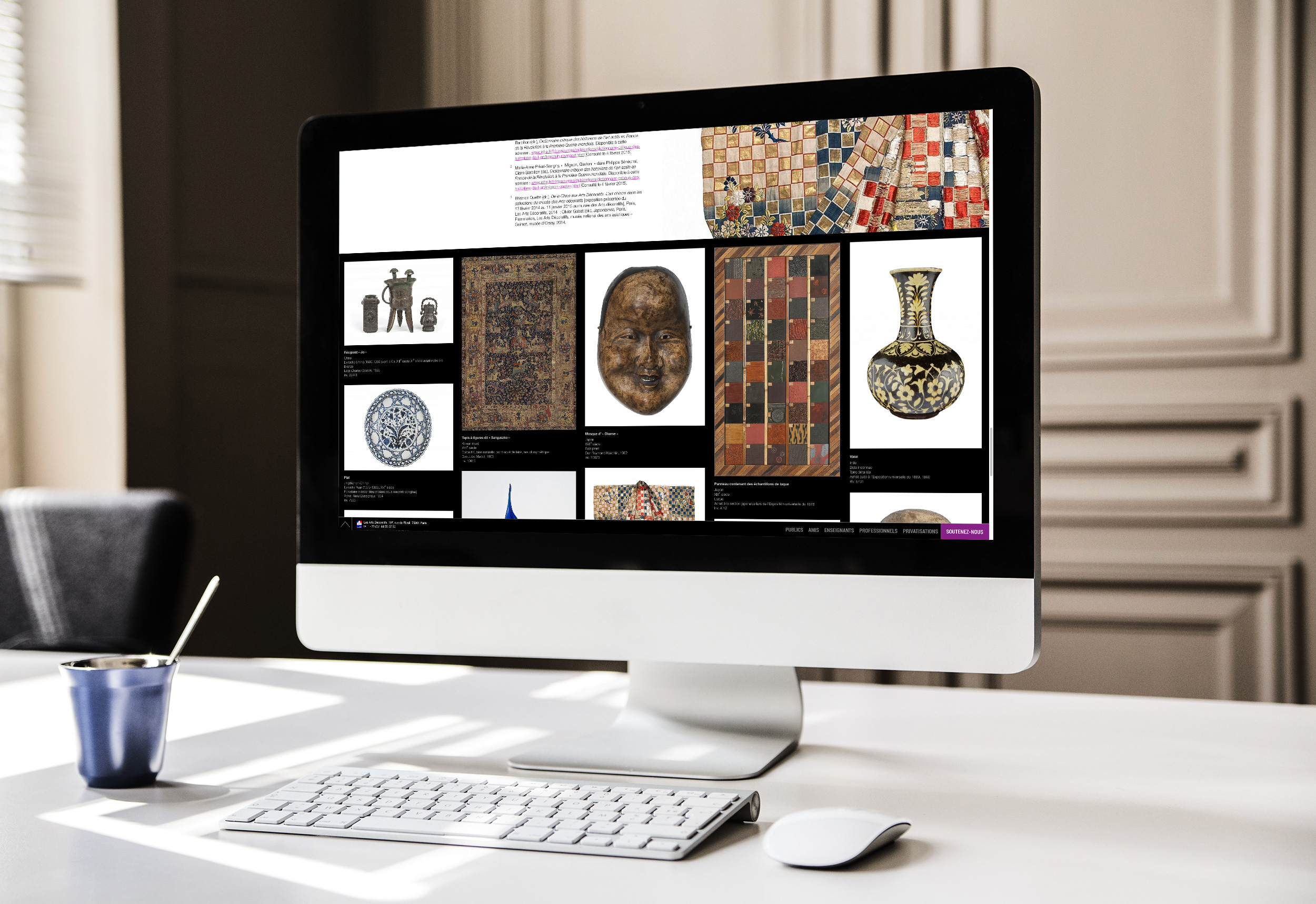 L'image montre un écran d'ordinateur affichant une collection d'objets artistiques ou archéologiques. Sur le fond noir, plusieurs éléments sont disposés : des motifs colorés, des sculptures, des vases et des artefacts ornementaux. Un ordinateur de bureau est posé sur un bureau minimaliste, avec un clavier et une petite tasse en dessous de l'écran. L'environnement semble moderne et épuré, avec un éclairage naturel provenant de la gauche.