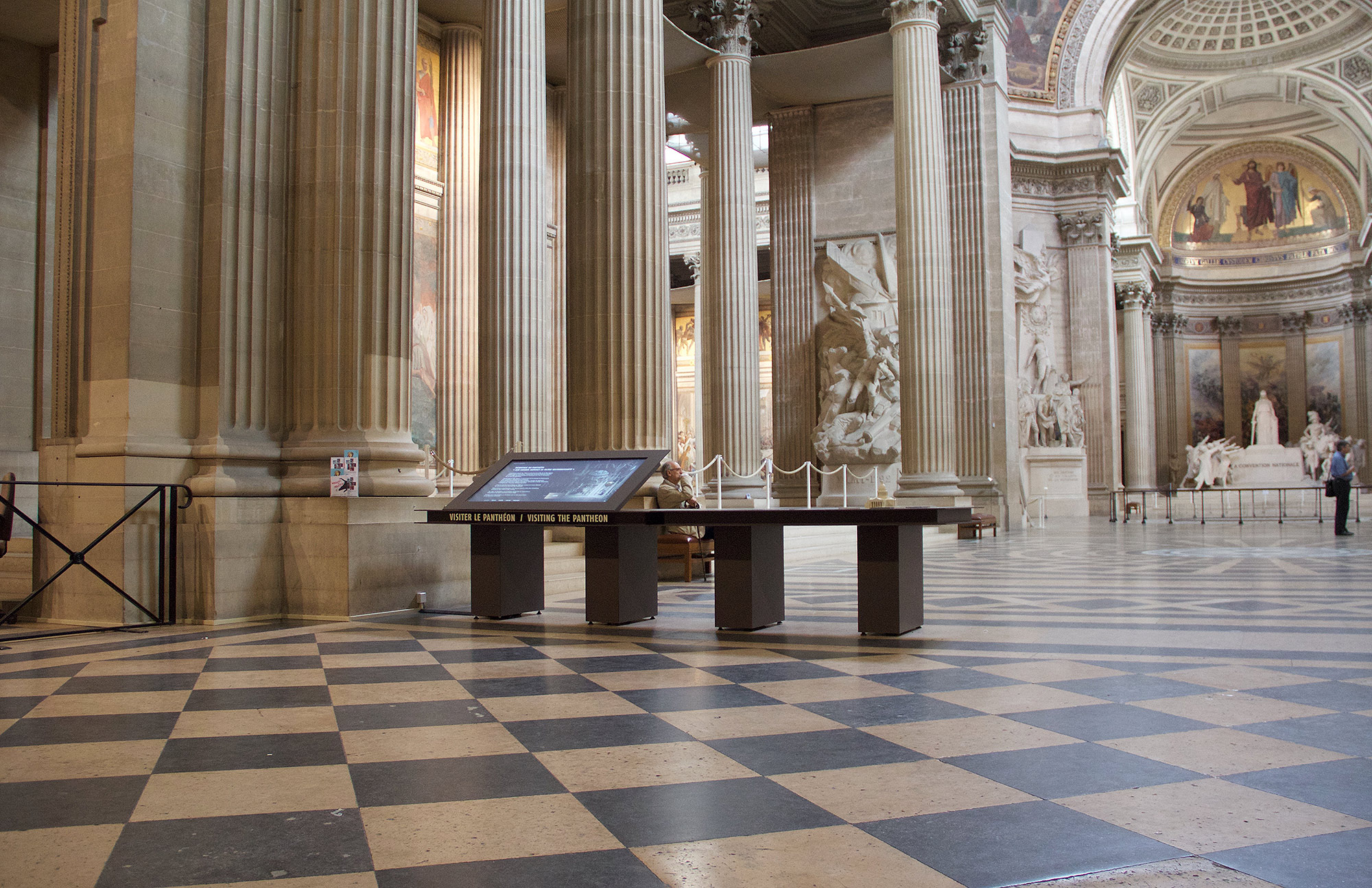 L'image montre l'intérieur d'un grand bâtiment, probablement un monument historique comme un panthéon. On peut observer de hauts plafonds ornés et des colonnes majestueuses. Le sol est décoré de carreaux en damier noir et blanc, créant un effet visuel intéressant. Au centre, il y a un écran interactif sur un socle, accompagné de barrières. En arrière-plan, on distingue des sculptures et des œuvres d'art sur les murs et une statue, ajoutant une atmosphère culturelle à l'espace.