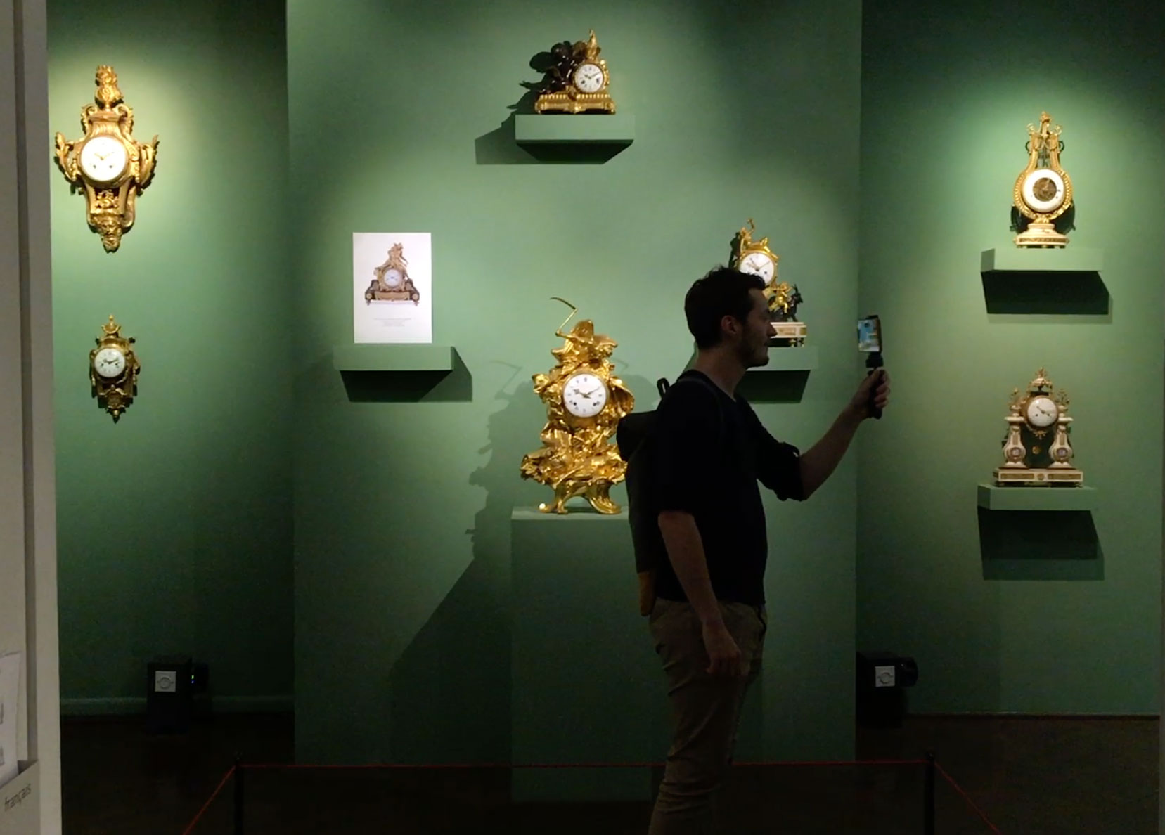 L'image montre une exposition dans un musée ou une galerie. Sur le mur vert, plusieurs horloges décoratives sont présentées sur des étagères. L'une des horloges est encadrée par une plaque explicative. Au premier plan, un homme tient un téléphone, probablement pour prendre des photos ou enregistrer des informations. L'éclairage met en valeur les détails des horloges, qui semblent être des pièces artistiques anciennes et ornées.