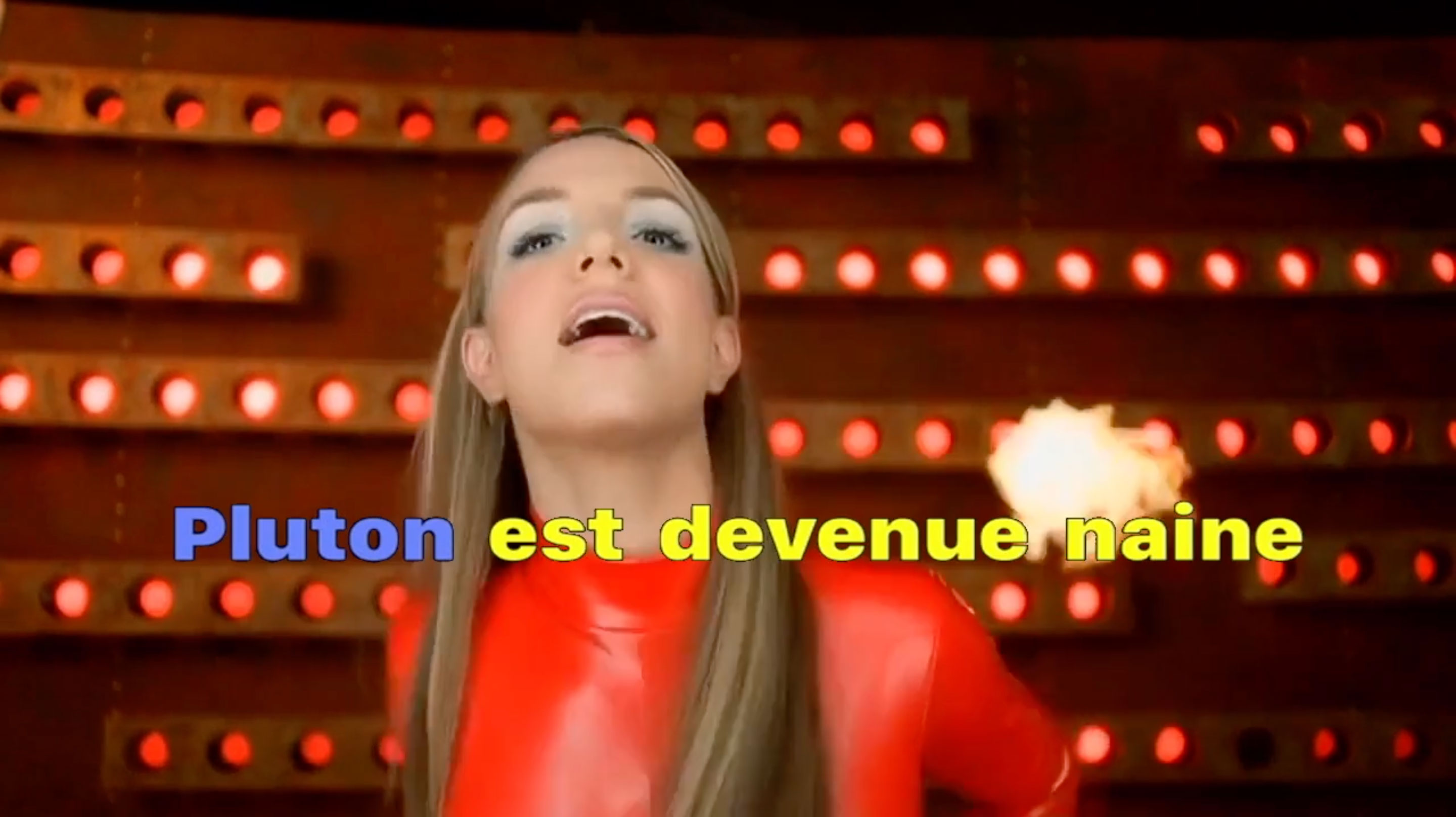 L'image montre une personne en train de chanter, probablement dans un cadre dynamique et coloré. Elle porte un costume rouge éclatant et a les cheveux longs. En arrière-plan, il y a des lumières clignotantes. Les mots « Pluton est devenue naine » sont affichés en grandes lettres colorées. Cela semble évoquer une thématique amusante ou ludique, peut-être liée à une chanson.