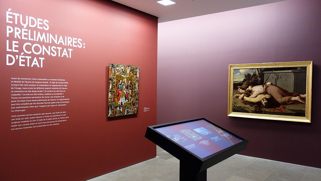 L'image montre une exposition dans un musée. Sur un mur d'une teinte rougeâtre, il y a un texte en blanc qui indique "ÉTUDES PRÉLIMINAIRES : LE CONSTAT D'ÉTAT". À côté, on peut voir une œuvre artistique encadrée, représentant une scène avec des figures humaines. Au premier plan, il y a un écran ou une table interactive qui semble contenir plus d'informations ou des illustrations liées à l'exposition. L'espace semble bien éclairé et organise une mise en valeur des œuvres présentées.