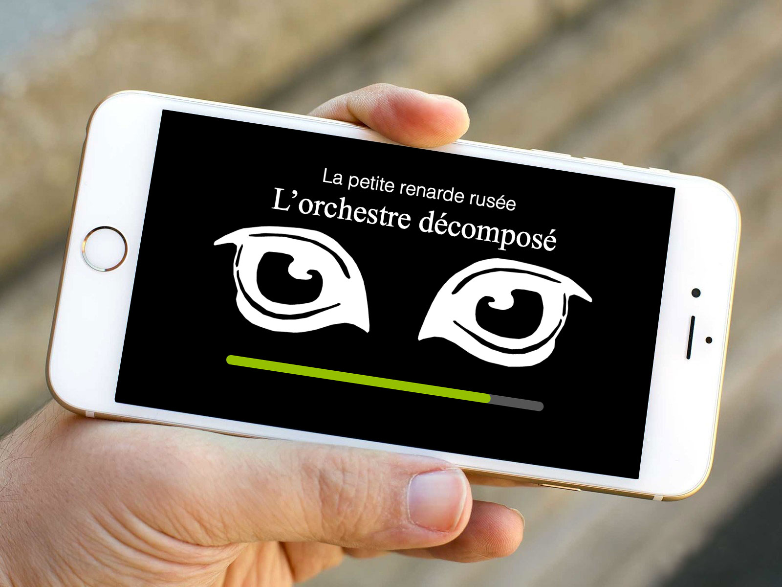 L'image montre un smartphone tenu dans une main. L'écran du téléphone affiche un fond noir avec deux yeux stylisés en blanc. Au-dessus des yeux, il y a un texte qui dit "La petite renarde rusée" et, en dessous, le titre "L'orchestre décomposé". Une barre de chargement verte est visible en bas de l'écran.