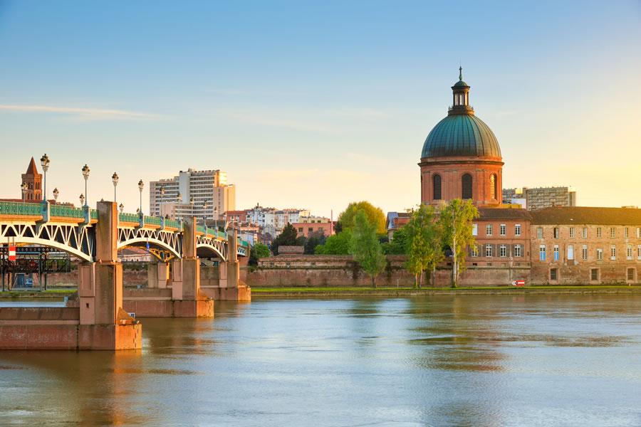 L'image montre un paysage urbain au bord d'une rivière, avec un pont qui enjambe l'eau. À droite, on aperçoit un bâtiment à la coupole verte, probablement une église ou un ancien bâtiment administratif. En arrière-plan, on distingue des immeubles modernes et des arbres qui ajoutent une touche de verdure. La lumière douce du soleil se reflète sur l'eau, créant une ambiance paisible et agréable. Ce cadre évoque une ville animée tout en offrant une vue sur la tranquillité de la rivière.
