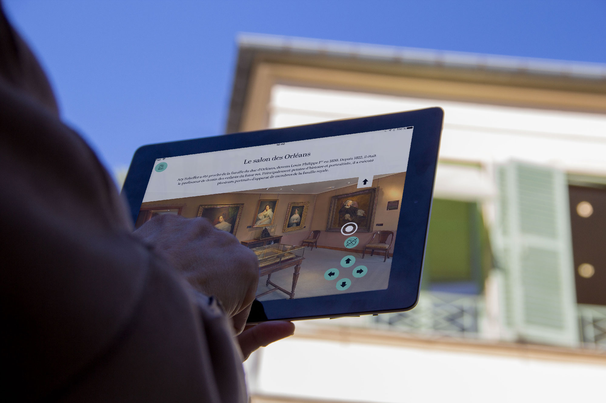 L'image montre une personne tenant une tablette dans un environnement extérieur, probablement près d'un musée ou d'une galerie d'art. Sur l'écran de la tablette, on peut voir une interface numérique qui semble présenter des informations sur une exposition, intitulée "Le salon des Oeuvres". En arrière-plan, il y a un bâtiment avec des volets ouverts et un ciel bleu, créant une ambiance agréable.