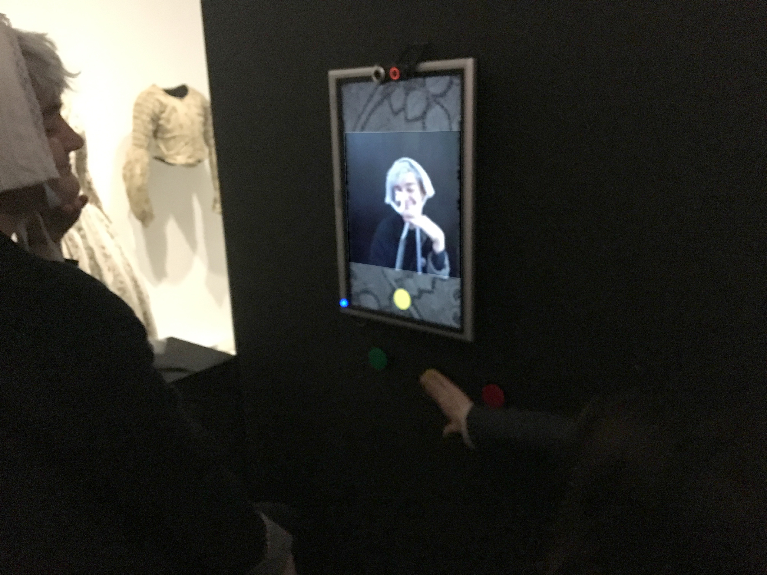 L'image montre une exposition où des visiteurs interagissent avec un écran. Sur l'écran, une personne semble réfléchir et regarder le spectateur. Il y a trois boutons colorés (rouge, jaune et vert) en dessous de l'écran, ce qui suggère une forme d'interaction, peut-être pour répondre à des questions ou choisir des options. Le fond est sombre, mettant en valeur l'écran et les visiteurs qui l'observent.