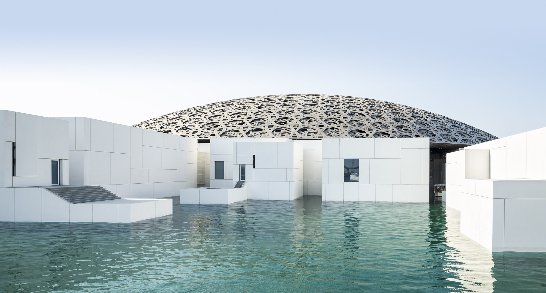 L'image montre une structure architecturale moderne qui semble être le Louvre Abu Dhabi. On peut voir une série de bâtiments blancs aux formes géométriques, entourés d'eau et sous un dôme distinctif en forme de grille. La surface de l'eau reflète la lumière, créant une ambiance paisible et contemporaine. Le ciel est clair et lumineux, ajoutant à l'élégance de l'ensemble.