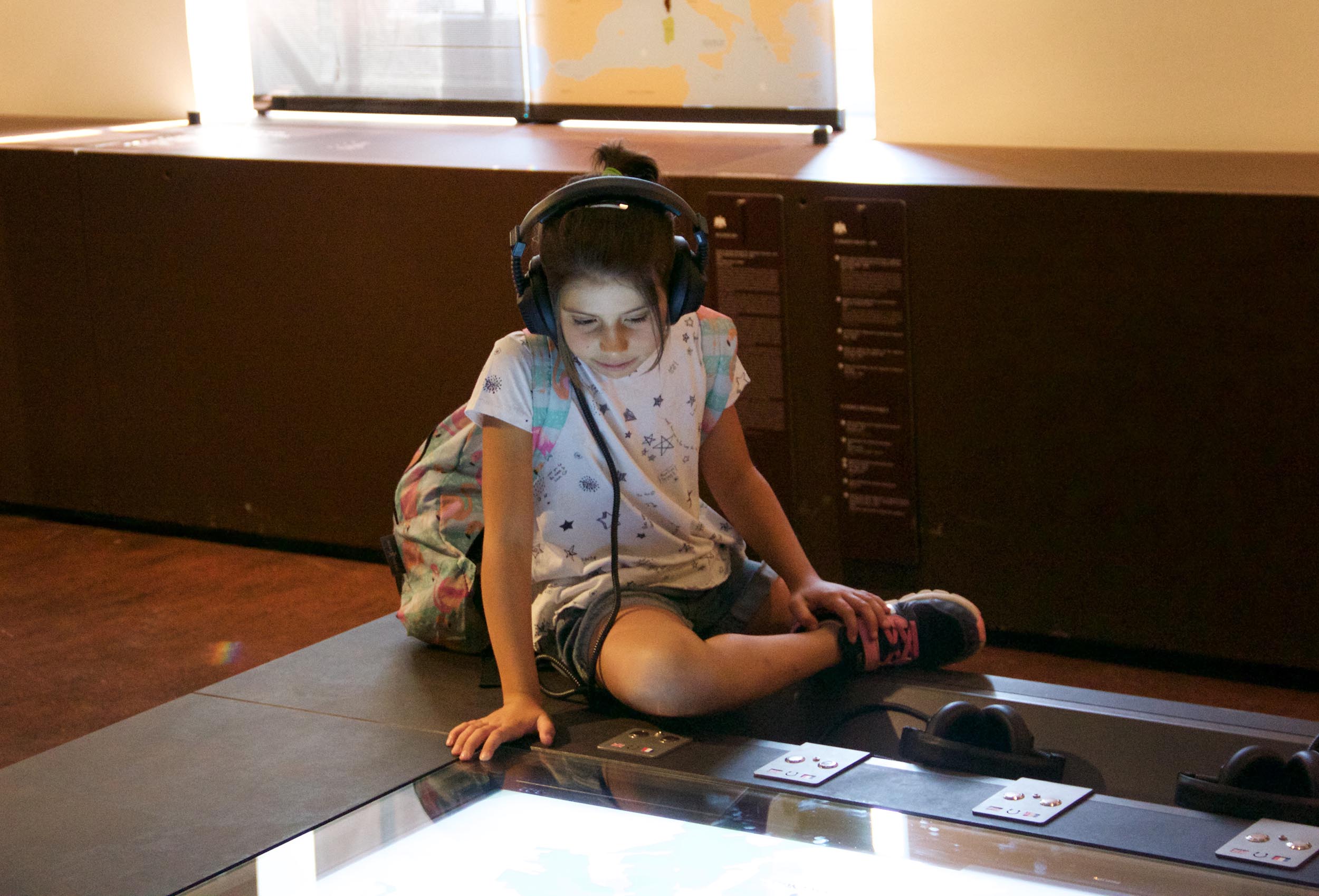 The image shows a young girl sitting on the floor at a museum or interactive exhibit. She is wearing headphones and appears to be focused on a touchscreen display in front of her. The girl has a backpack beside her and is dressed in casual clothing, including a T-shirt with a star pattern and shorts. The atmosphere looks educational, with various informational panels in the background.
