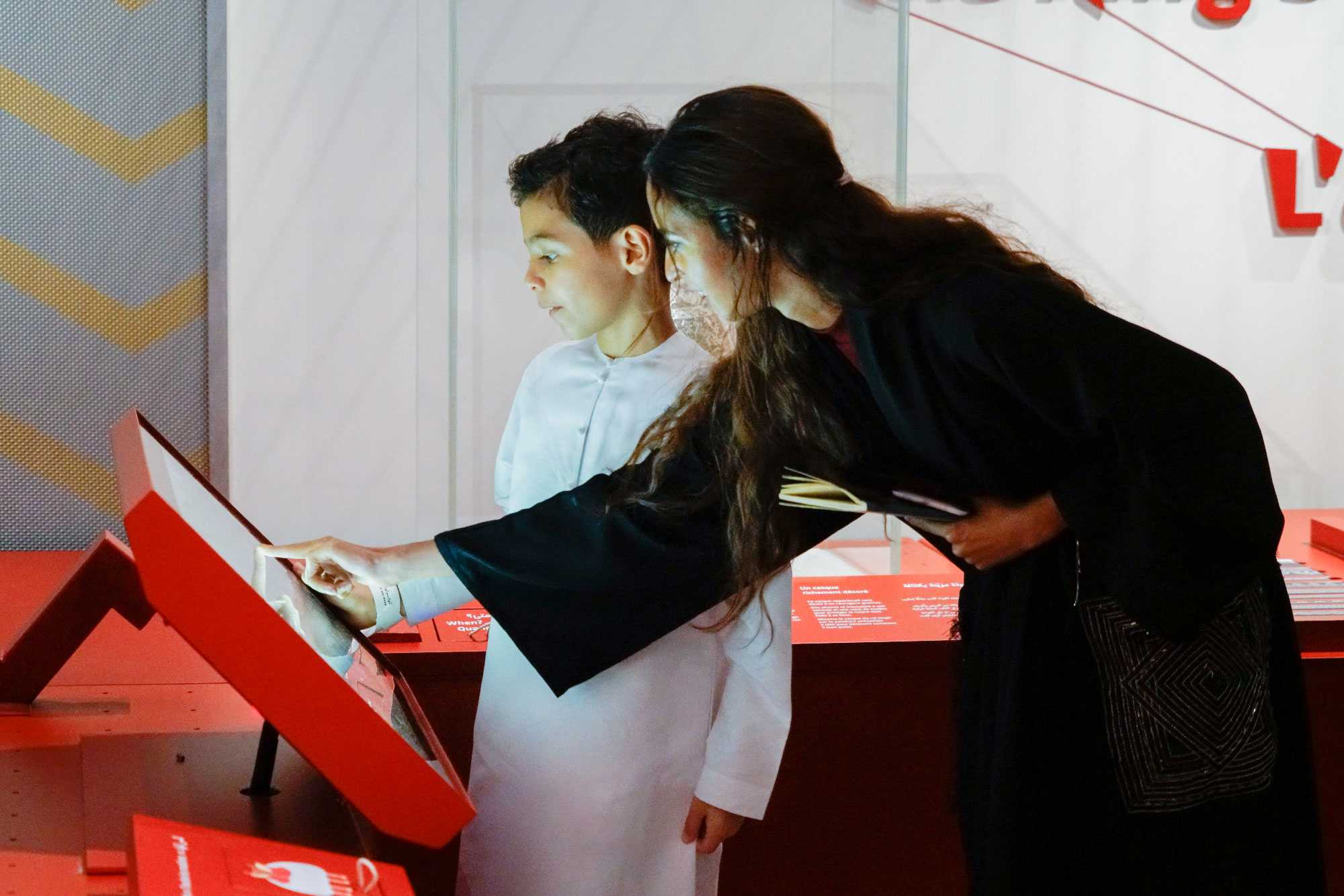 The image shows a young boy and a woman interacting with a display at an exhibition. The boy, wearing a white traditional outfit, is closely observing the screen while the woman, dressed in a dark garment, points to something on the display. Both appear engaged and curious as they explore the exhibit. The environment is modern, with red and white colors dominating the design.