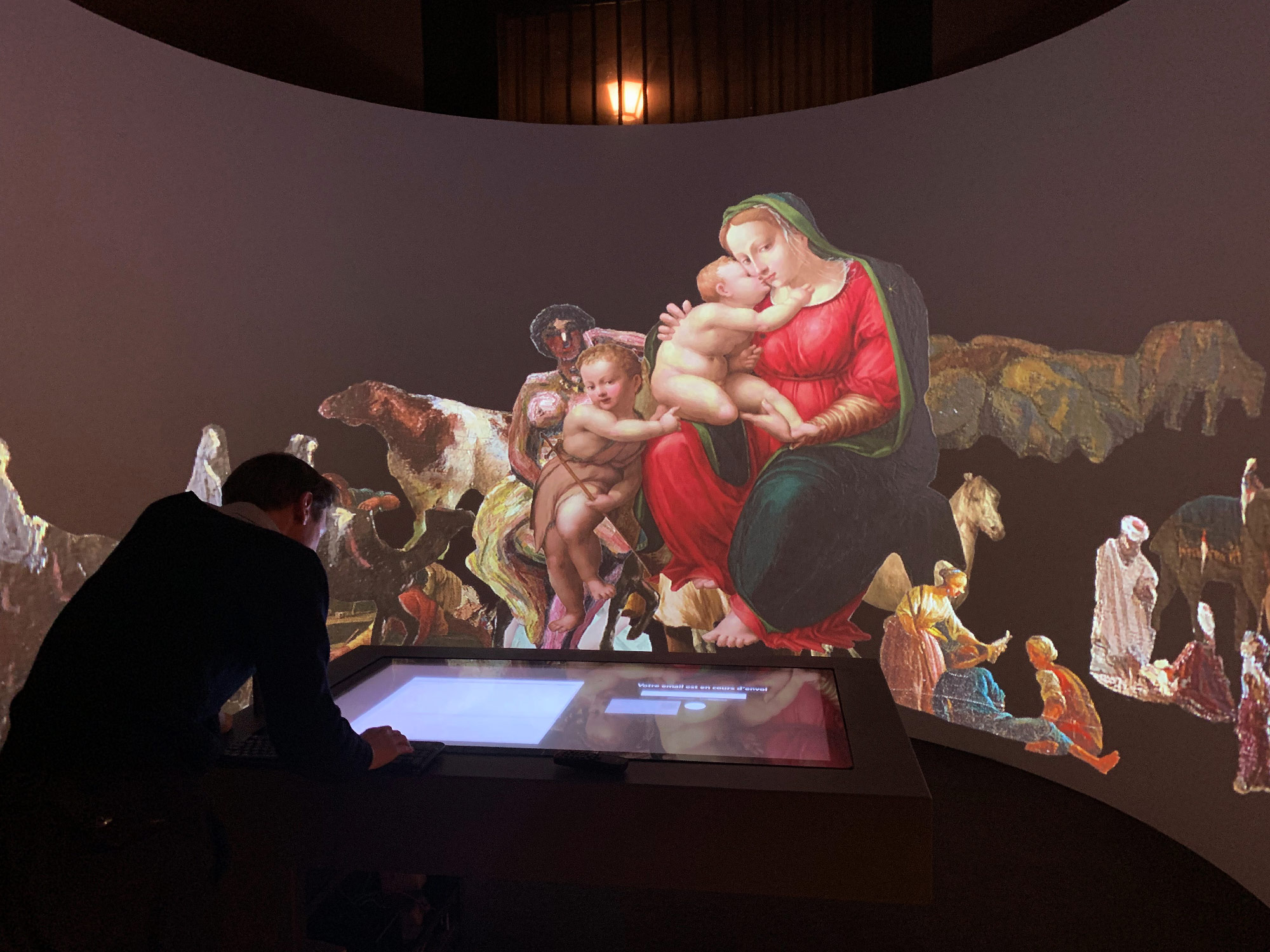 L'image montre une installation artistique immersive où une grande reproduction d'une œuvre d'art classique est projetée sur un mur. Un homme est penché sur une table interactive, semblant interagir avec l'exposition. En arrière-plan, des figures et des scènes colorées sont affichées, créant une atmosphère dynamique. Les éléments de l'œuvre incluent une Vierge à l'Enfant entourée de diverses figures humaines et animales, le tout dans un contexte artistique riche et captivant.