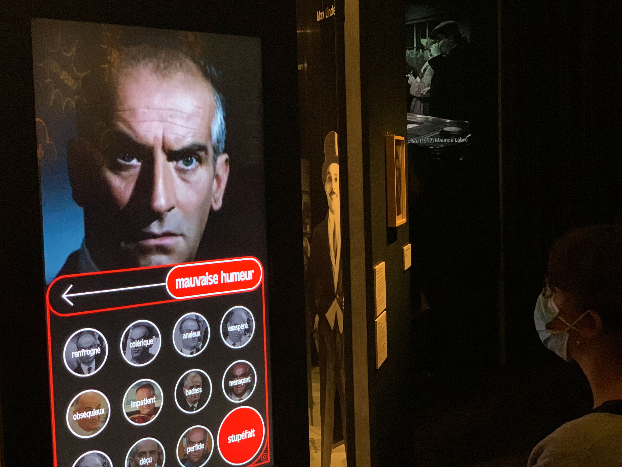 L'image montre une installation muséale avec un homme dont le visage est affiché sur un écran. Il regarde l'objectif avec une expression sérieuse. En bas de l'écran, il y a des mots qui semblent décrire différentes humeurs. Une personne portant un masque observe l'écran, suggérant une interaction avec l'affichage. À l'arrière-plan, on peut voir d'autres images en noir et blanc, ajoutant une atmosphère historique à l'ensemble de l'installation.