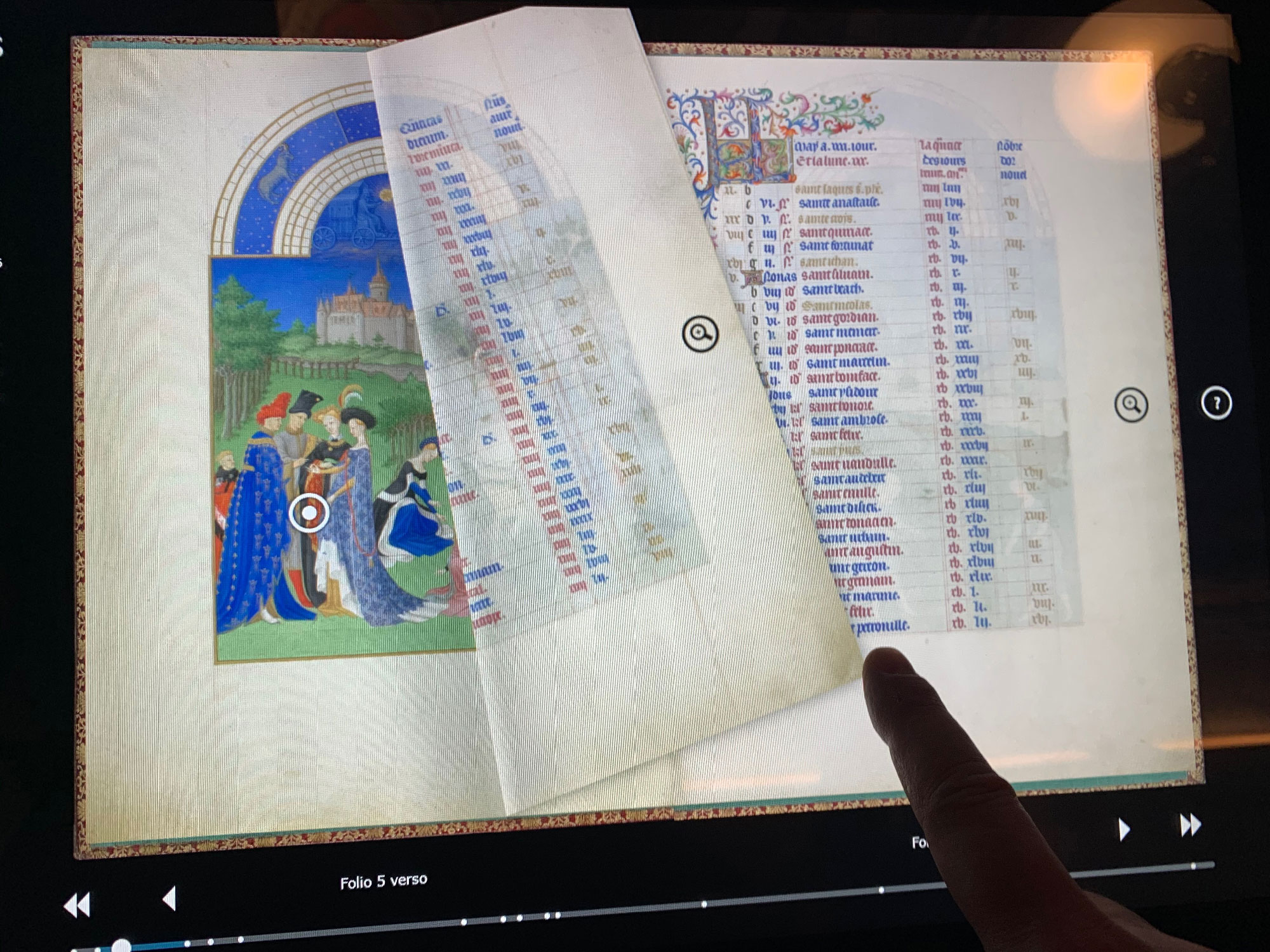 L'image montre une page d'un manuscrit ancien, probablement un livre d'heures ou un autre type de recueil médiéval. À gauche, on peut apercevoir une illustration colorée représentant des figures humaines vêtues de costumes d'époque, se tenant devant un paysage avec un château. À droite, une colonne de texte est visible, répertoriant probablement des noms ou des prières, accompagnée d'une décoration élaborée en haut. Un doigt pointe vers la page, suggérant une interaction avec le contenu. Le fond est sobre, mettant l'accent sur le manuscrit.