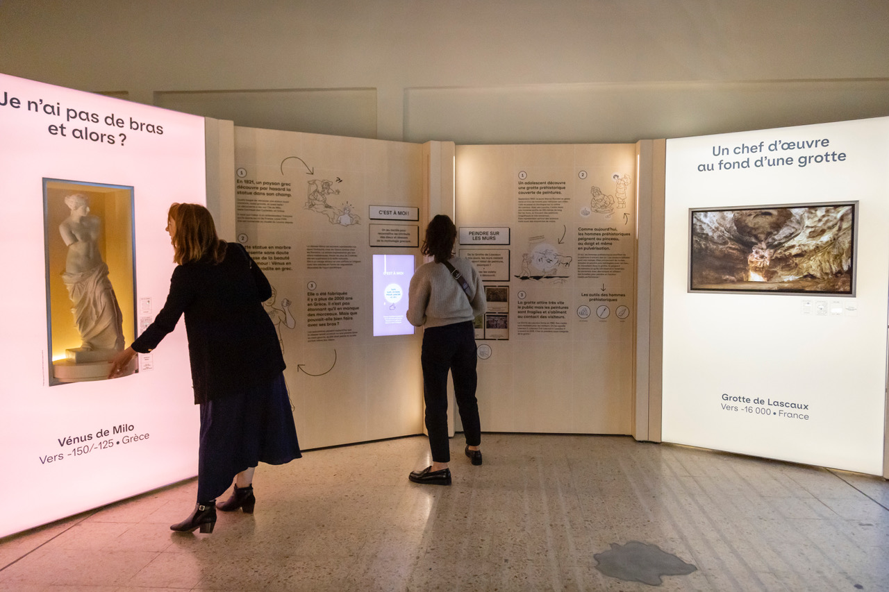 L'image montre une exposition muséale où deux personnes interagissent avec des installations. À gauche, une femme examine une vitrine qui semble contenir une statue. À droite, un panneau d'affichage présente des informations sur des œuvres d'art, incluant une mention de la "Vénus de Milo" et des grottes de Lascaux. L'espace est éclairé avec des lumières douces et des éléments graphiques qui attirent l'attention des visiteurs.
