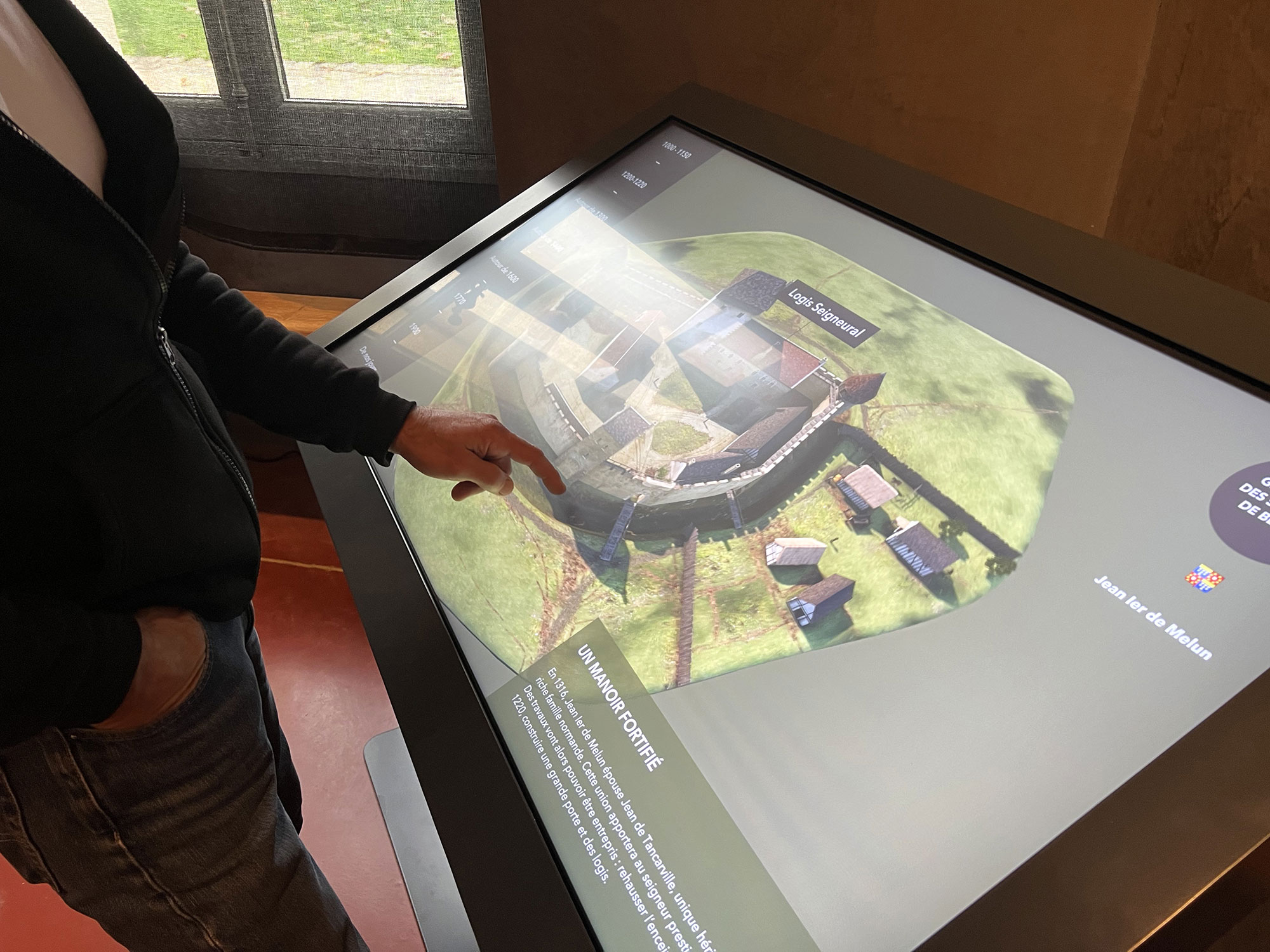 L'image montre une personne en train d'interagir avec un écran tactile. L'écran affiche une vue en 3D d'une fortification ou d'un site historique, avec des bâtiments et un espace fortifié bien définis. L'utilisateur semble examiner les détails de la présentation, en utilisant son doigt pour pointer ou interagir avec certaines parties de l'image. En arrière-plan, on peut voir des fenêtres qui laissent passer la lumière naturelle, créant une atmosphère accueillante.