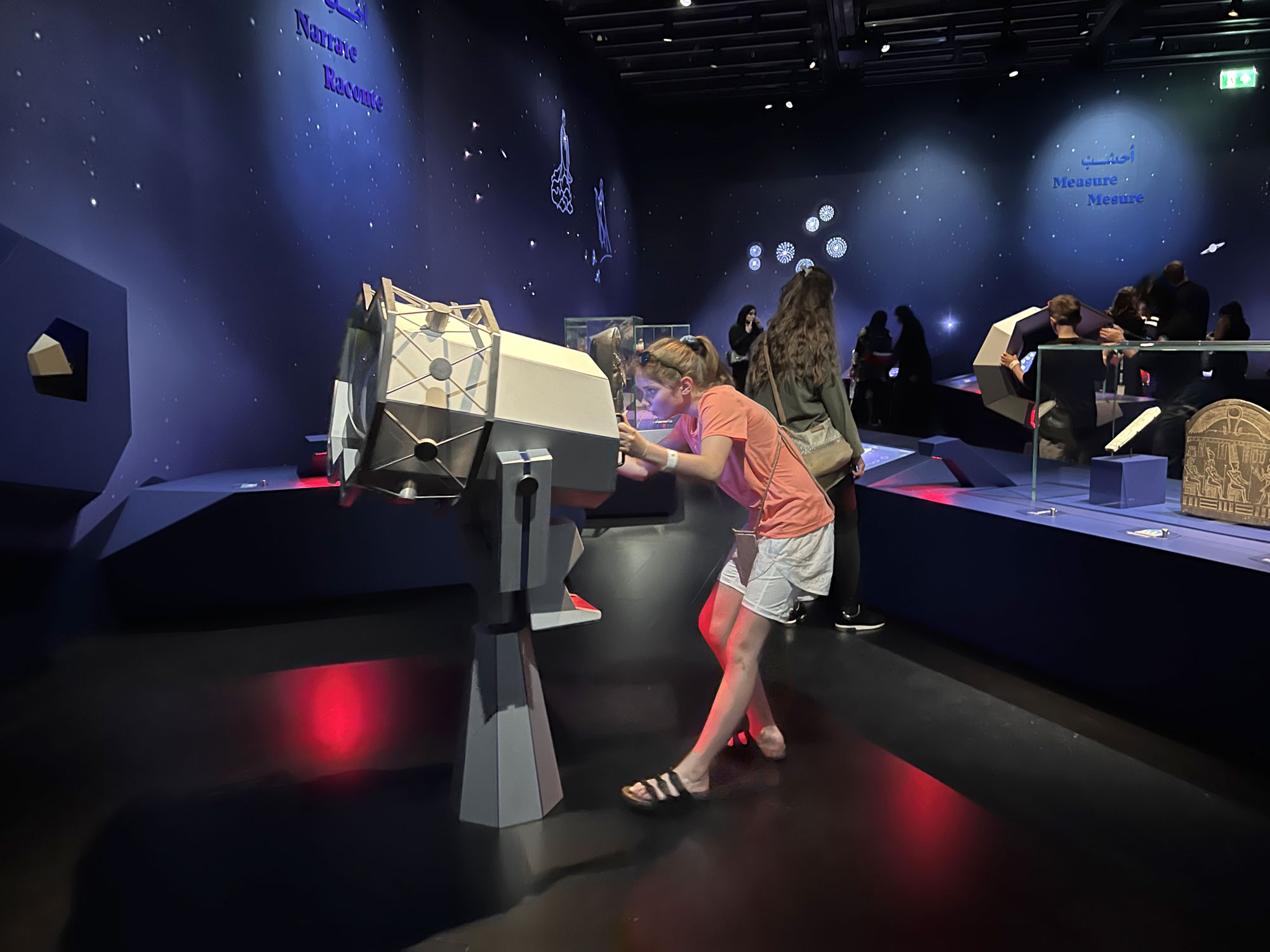 L'image montre une exposition dans un musée ou un espace éducatif. Une jeune fille se penche sur un appareil interactif ou une installation en forme de cylindre. Elle semble curieuse, examinant l'objet de près. En arrière-plan, plusieurs visiteurs explorent l'exposition, tandis que des éléments d'affichage et des décorations murales évoquent des thèmes scientifiques ou astronomiques. L'éclairage est doux, créant une ambiance agréable.