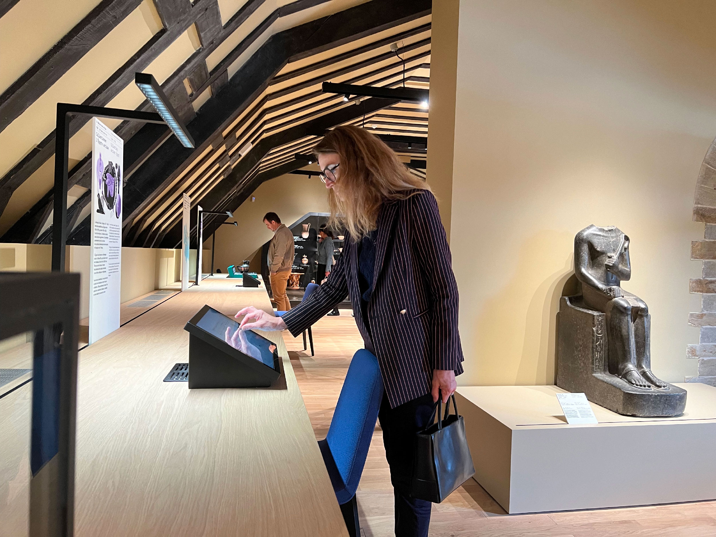 L'image montre une femme se tenant près d'un comptoir dans un musée. Elle semble interagir avec un écran tactile. En arrière-plan, on peut apercevoir d'autres visiteurs et une statue ou une sculpture ancienne sur un socle. La pièce a des poutres en bois au plafond, créant une ambiance chaleureuse et accueillante. La femme porte une tenue professionnelle avec un sac à main.