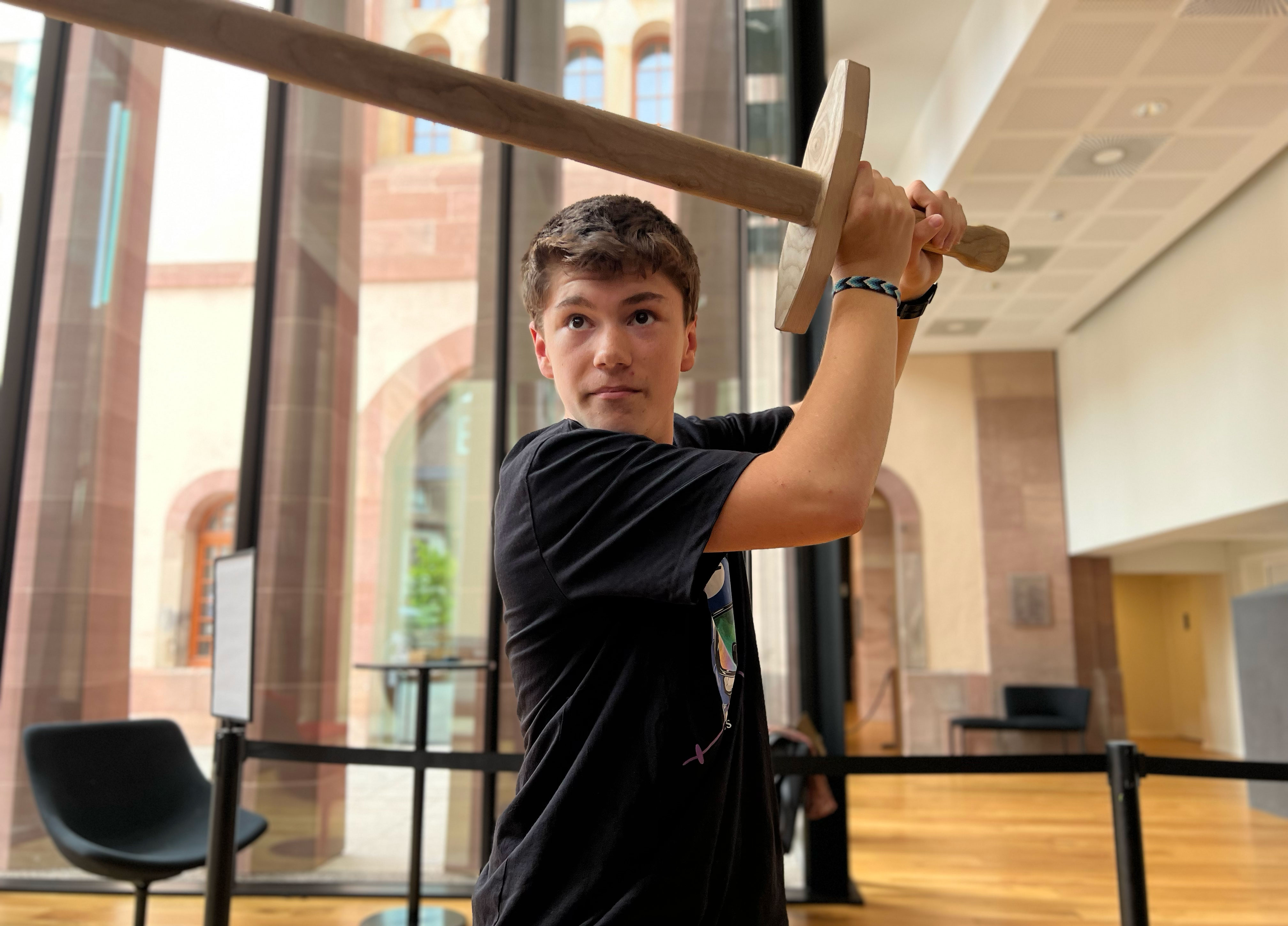 L'image montre un jeune homme levant une épée en bois. Il se tient dans un espace intérieur, probablement dans un musée ou un bâtiment historique, avec de grandes fenêtres et un design moderne. Le jeune homme porte un t-shirt noir et a une expression concentrée, semblant prêt à se battre ou à s'entraîner. L'ambiance générale est dynamique et engageante.