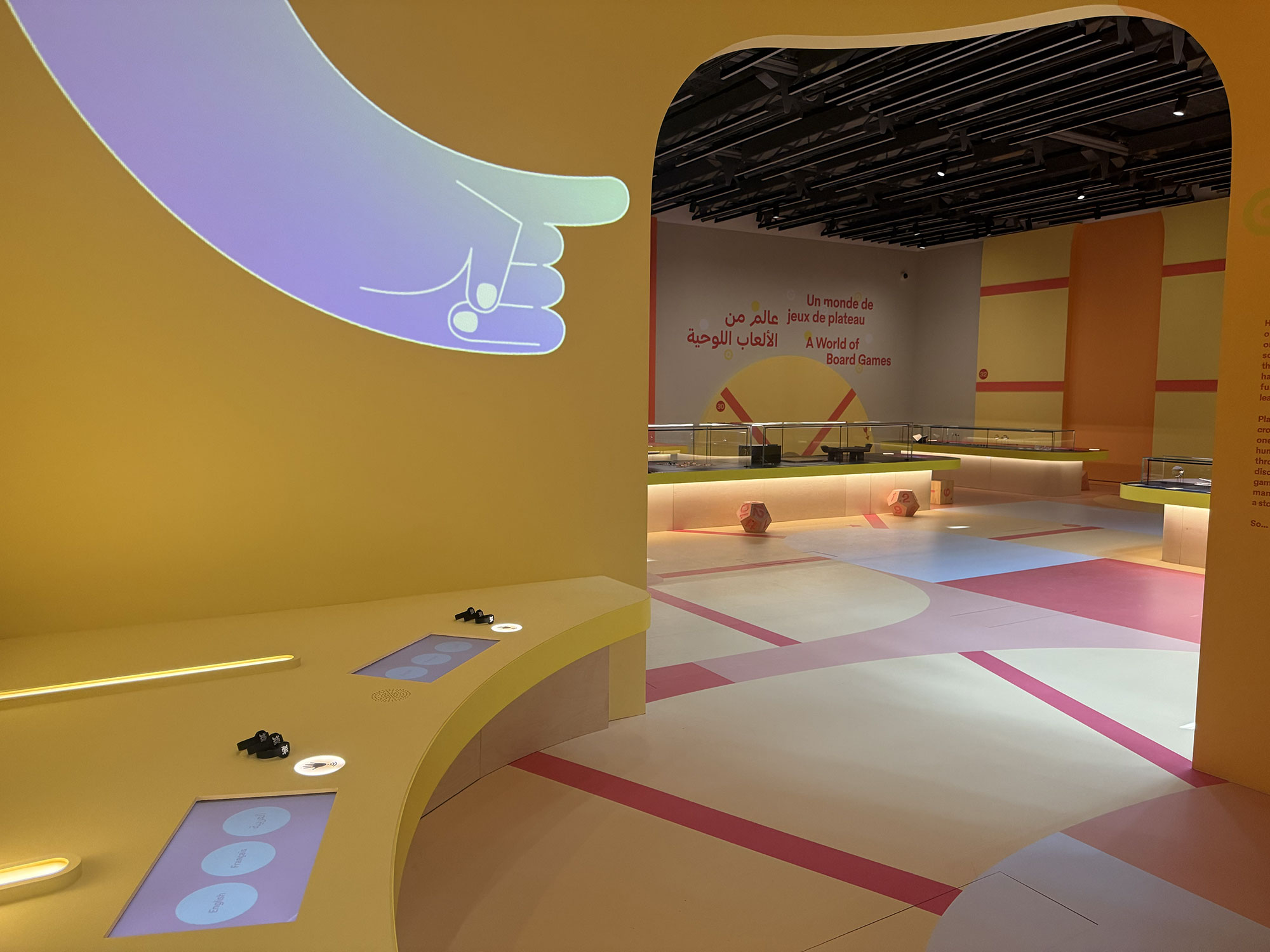L'image montre un espace intérieur coloré avec des murs jaunes et des éléments de design modernes. On peut voir des tables avec des dispositifs interactifs et de l'éclairage doux. À l'arrière-plan, il y a des inscriptions en arabe et en français, indiquant une exposition sur les jeux de société. L'agencement de la pièce semble accueillant et ludique, avec des tapis de sol aux couleurs variées, créant une atmosphère conviviale.