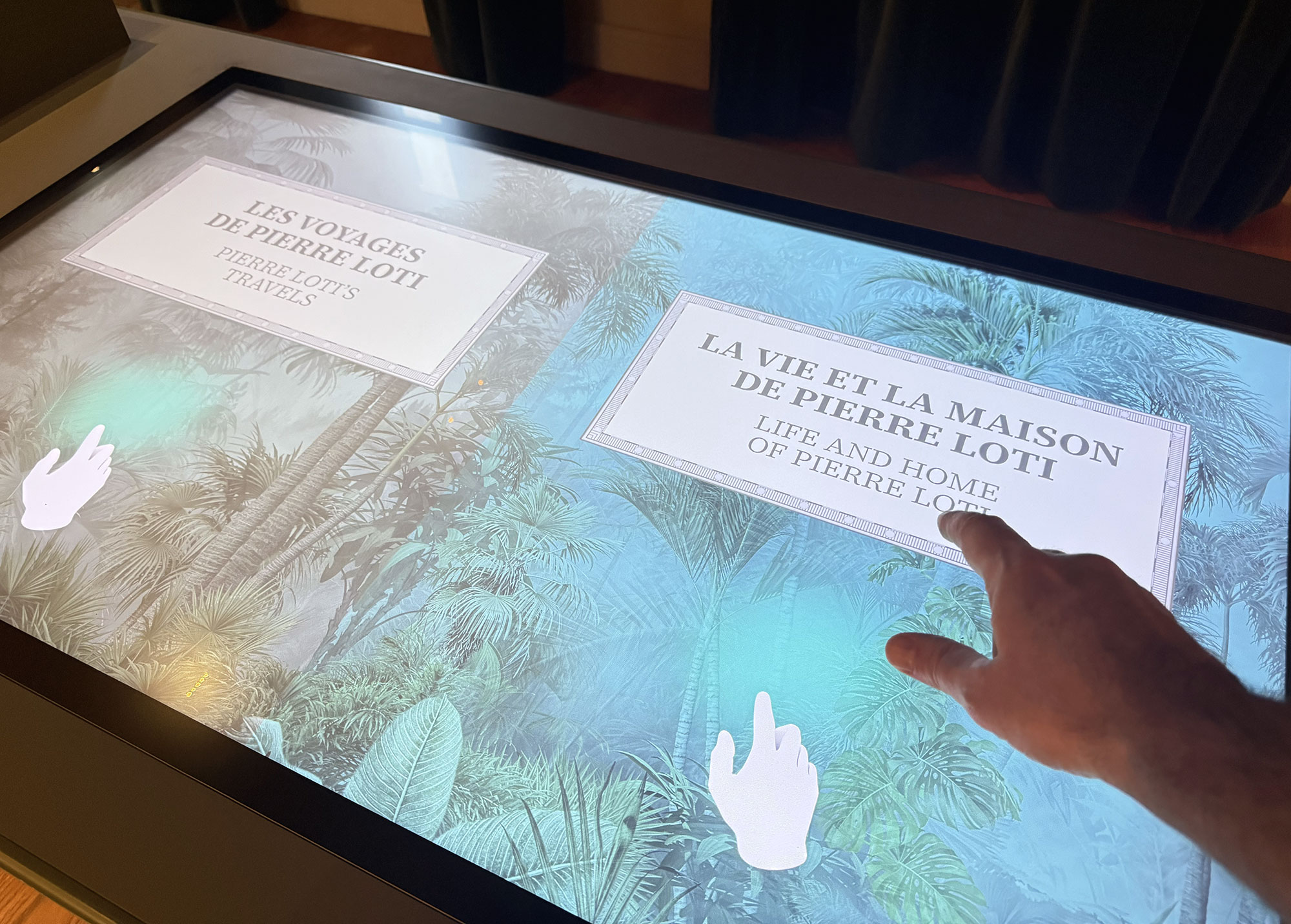 L'image montre un écran tactile interactif avec deux sections principales. À gauche, on peut lire "Les voyages de Pierre Loti", et à droite, "La vie et la maison de Pierre Loti", en français et en anglais. En bas, une main pointe vers l'écran, suggérant une interaction. L'arrière-plan est une illustration de paysages tropicaux, évoquant probablement les thèmes des voyages de Pierre Loti.