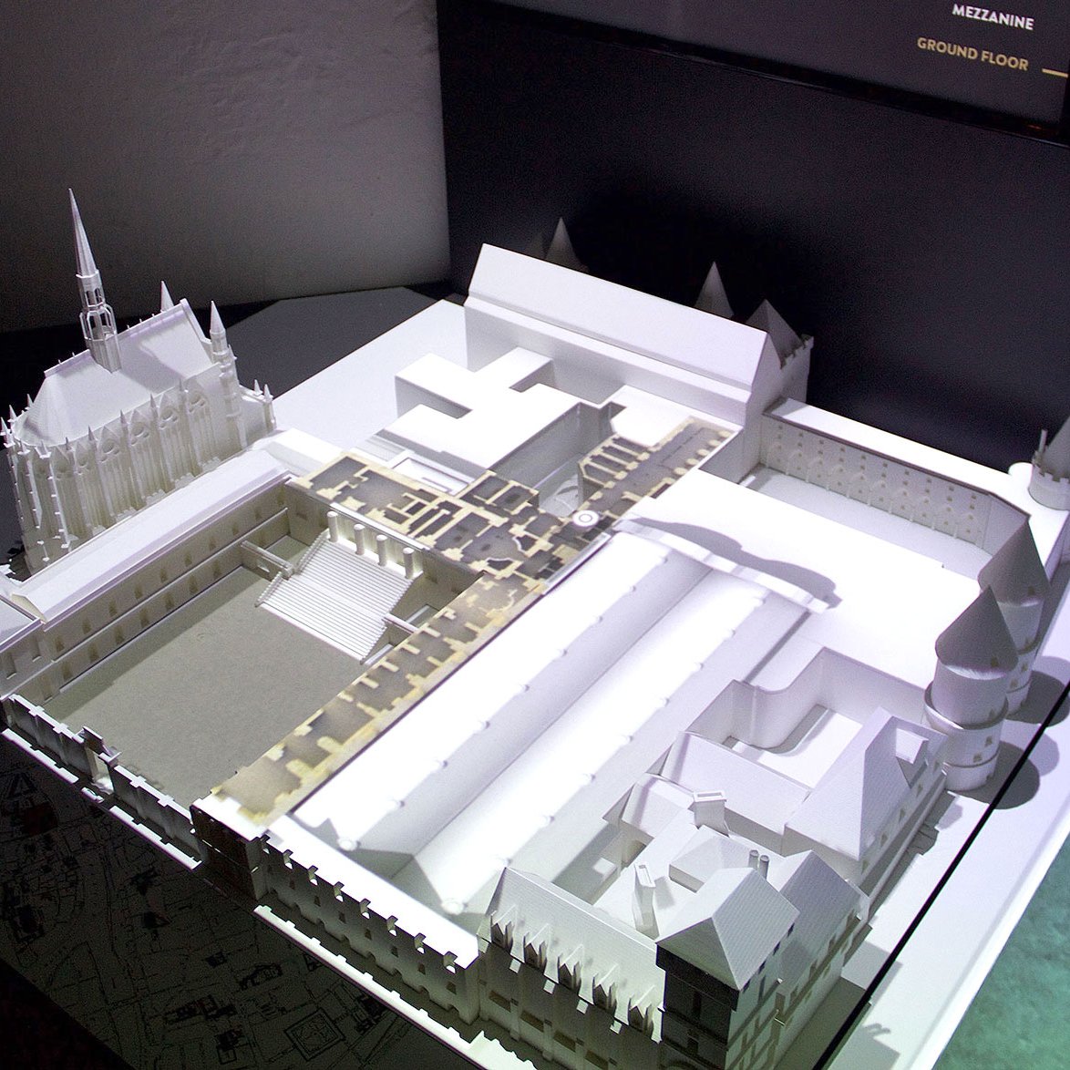 L'image montre une maquette architecturale en trois dimensions, probablement d'un bâtiment ou d'un site historique. La maquette est éclairée, mettant en évidence divers détails, comme les formes des bâtiments, les toits et les espaces environnants. On peut distinguer des structures avec des éléments architecturaux variés, et il semble qu'il y ait une partie en hauteur, probablement une tour. L'illustration permet de visualiser l'agencement des lieux, offrant un aperçu de l'édifice lorsqu'il était à l'état original.