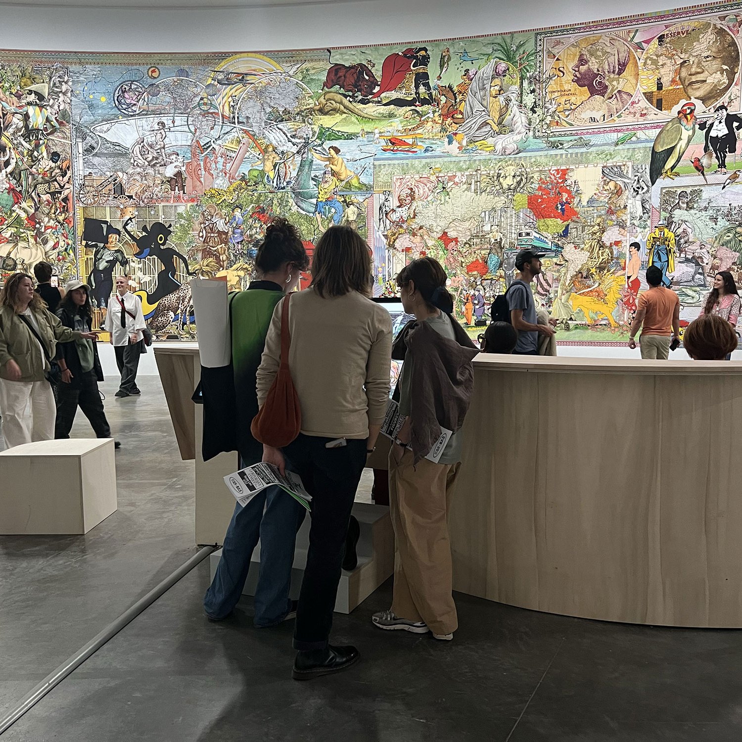 L'image montre une galerie d'art où plusieurs personnes explorent une œuvre murale colorée qui occupe toute la surface d'un mur. La scène est animée, avec des visiteurs discutant et observant l'art. Les personnages sur le mur semblent provenir de diverses influences artistiques et culturelles, créant une atmosphère dynamique et engageante. Des structures en bois, probablement des présentoirs ou des zones de repos, sont visibles au premier plan.