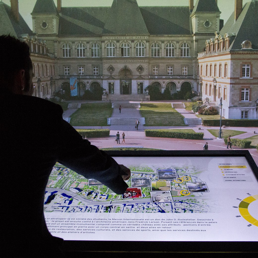 L'image montre une personne, probablement un homme, se tenant devant un grand écran qui affiche une vue de la place autour d'un bâtiment historique. Il interagit avec une carte affichée sur une surface tactile, pointant du doigt un endroit spécifique. Le fond révèle une architecture classique avec des détails élégants et une atmosphère de découverte urbaine. L'ensemble donne une impression de modernité alliée à l'histoire, comme une exposition ou un projet d'aménagement.