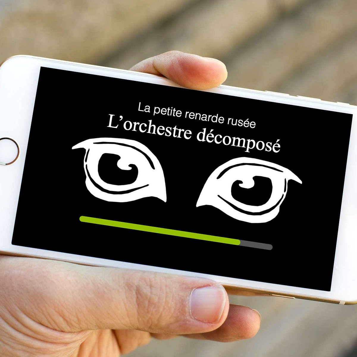L'image montre un smartphone tenu dans une main. L'écran du téléphone affiche un fond noir avec deux yeux stylisés en blanc. Au-dessus des yeux, il y a un texte qui dit "La petite renarde rusée" et, en dessous, le titre "L'orchestre décomposé". Une barre de chargement verte est visible en bas de l'écran.