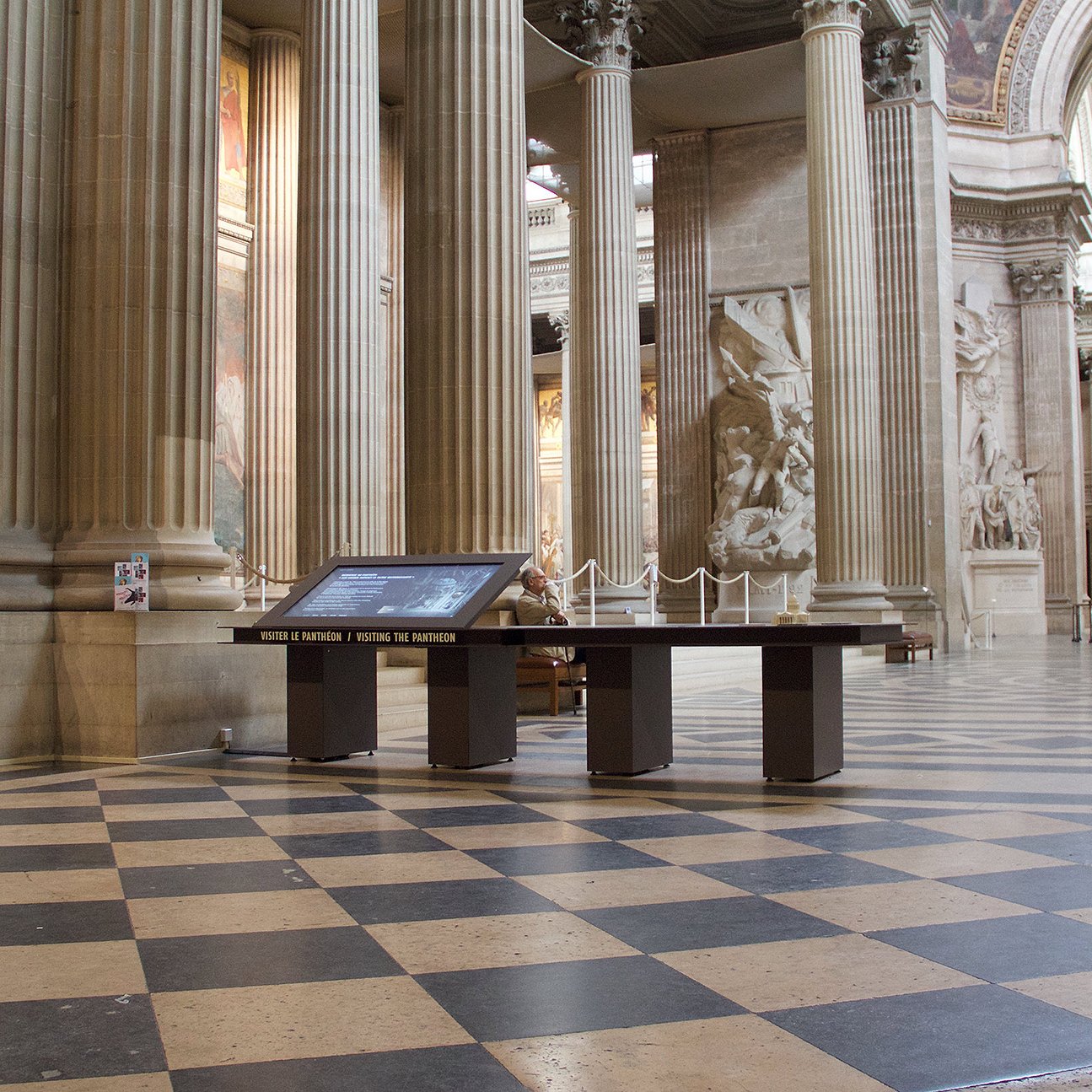 L'image montre l'intérieur d'un grand bâtiment, probablement un monument historique comme un panthéon. On peut observer de hauts plafonds ornés et des colonnes majestueuses. Le sol est décoré de carreaux en damier noir et blanc, créant un effet visuel intéressant. Au centre, il y a un écran interactif sur un socle, accompagné de barrières. En arrière-plan, on distingue des sculptures et des œuvres d'art sur les murs et une statue, ajoutant une atmosphère culturelle à l'espace.