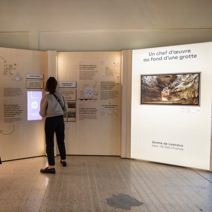 L'image montre une exposition muséale où deux personnes interagissent avec des installations. À gauche, une femme examine une vitrine qui semble contenir une statue. À droite, un panneau d'affichage présente des informations sur des œuvres d'art, incluant une mention de la "Vénus de Milo" et des grottes de Lascaux. L'espace est éclairé avec des lumières douces et des éléments graphiques qui attirent l'attention des visiteurs.